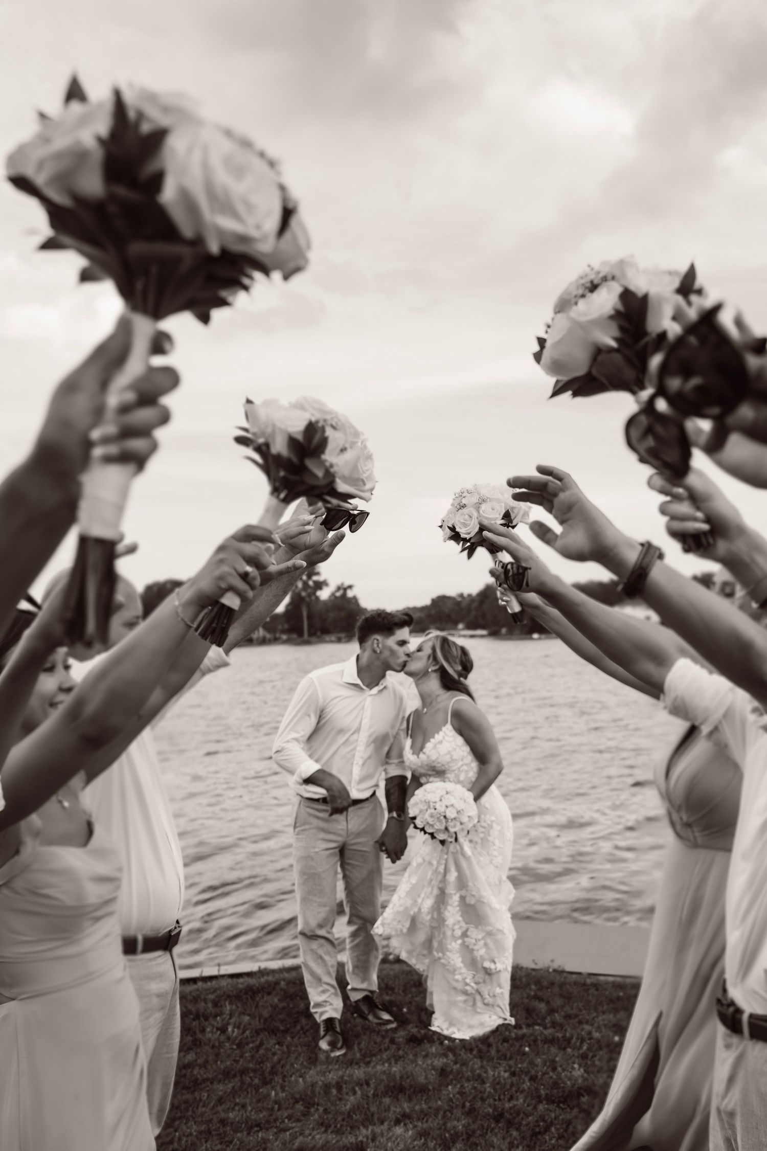 Wedding party holds bouquets aloft while a couple kisses on a waterfront lawn in black and white photography.