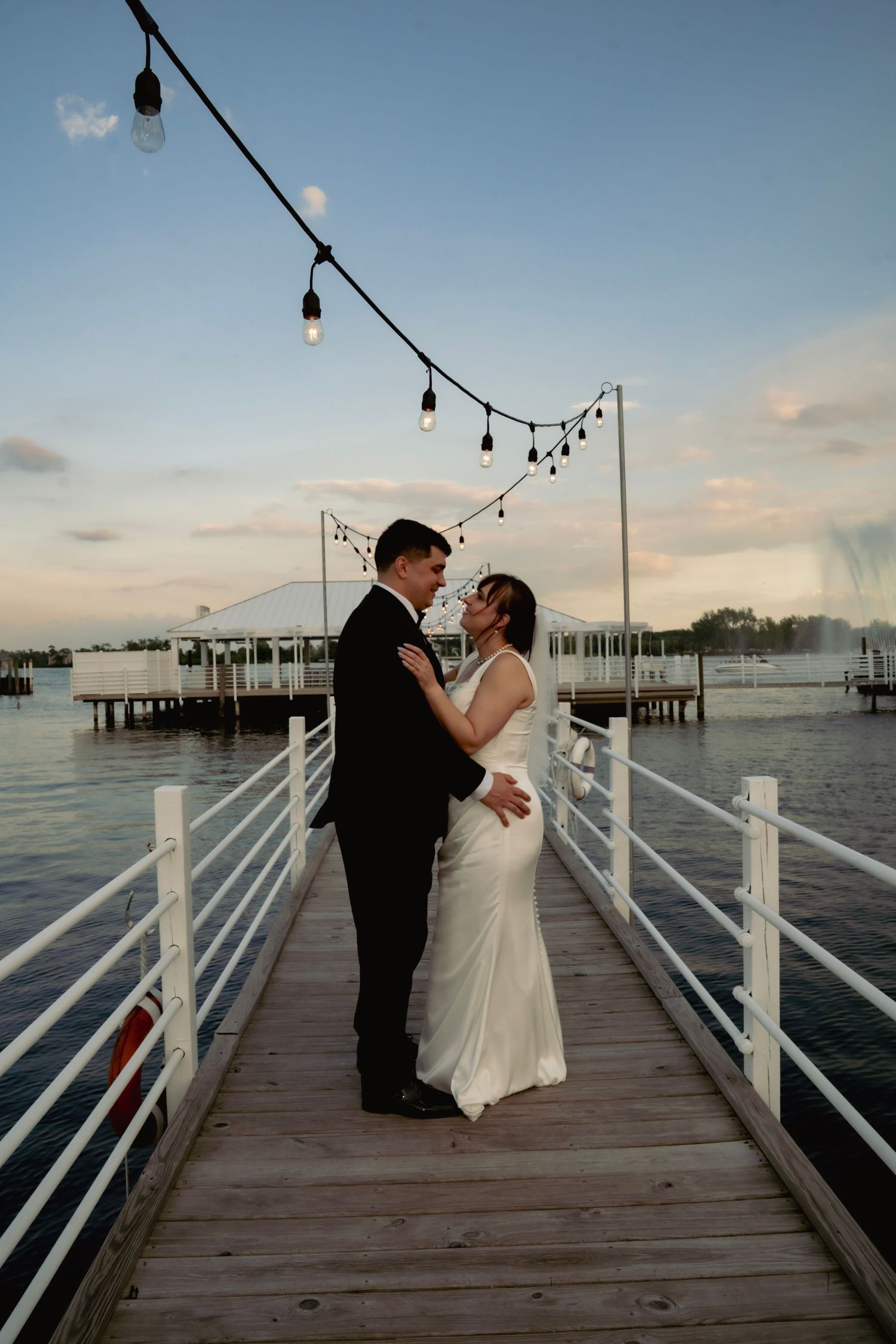 A romantic couple shares their first dance at sunset on a wooden pier decorated with string lights overlooking the water.