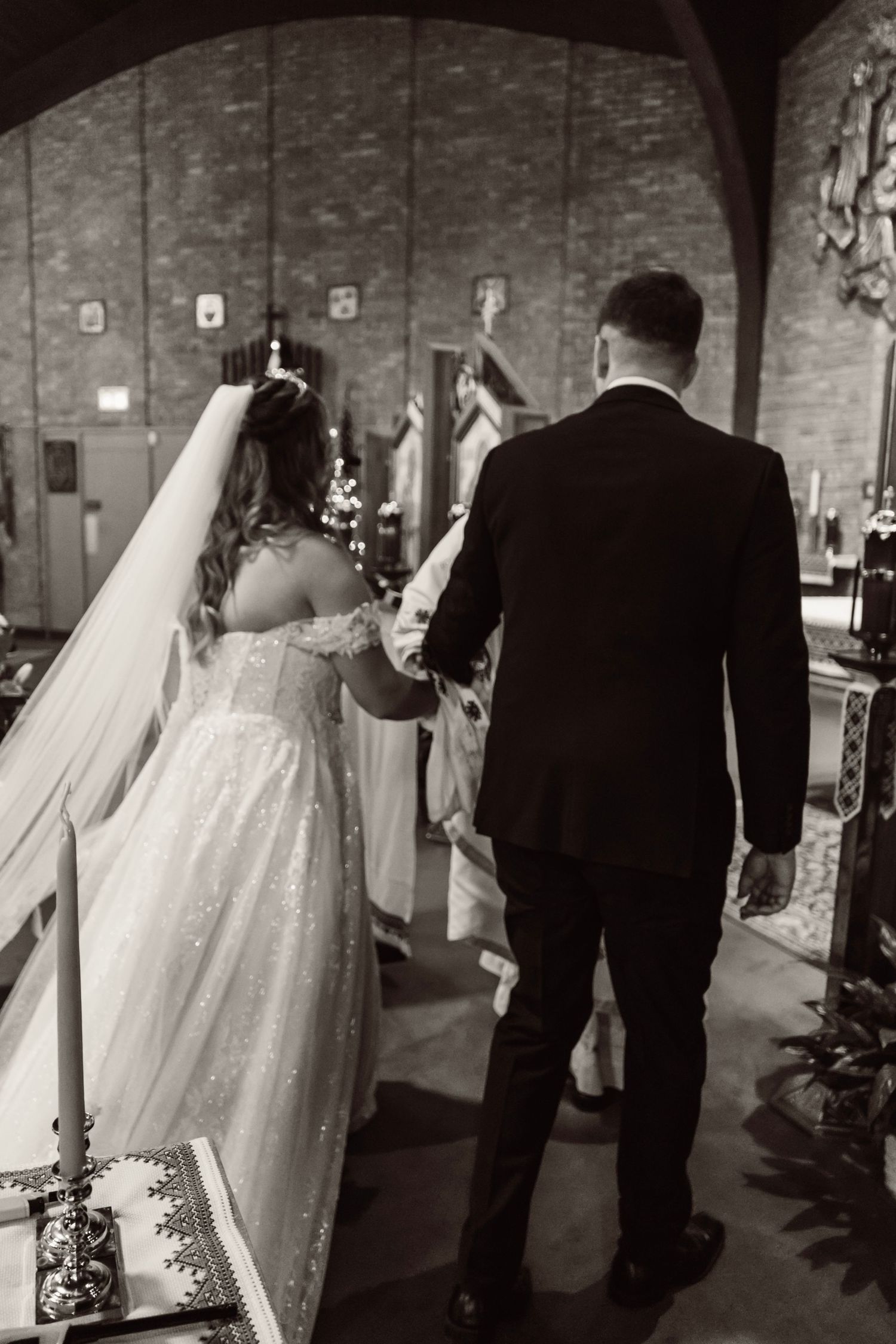 A black and white photo of newlyweds walking together through an arched brick venue with string lights overhead.