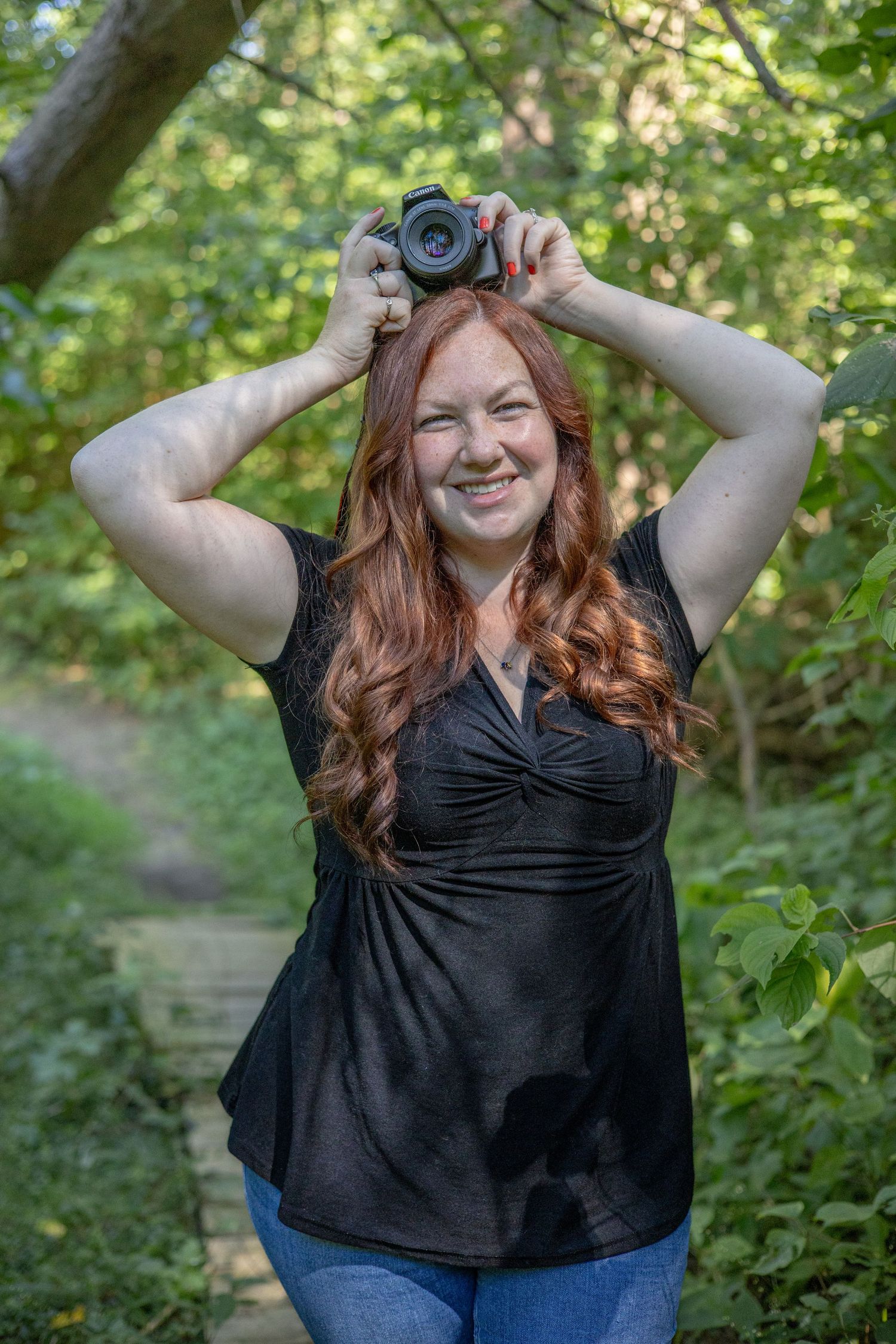 A photographer with long red hair playfully holding up a camera in a wooded setting.