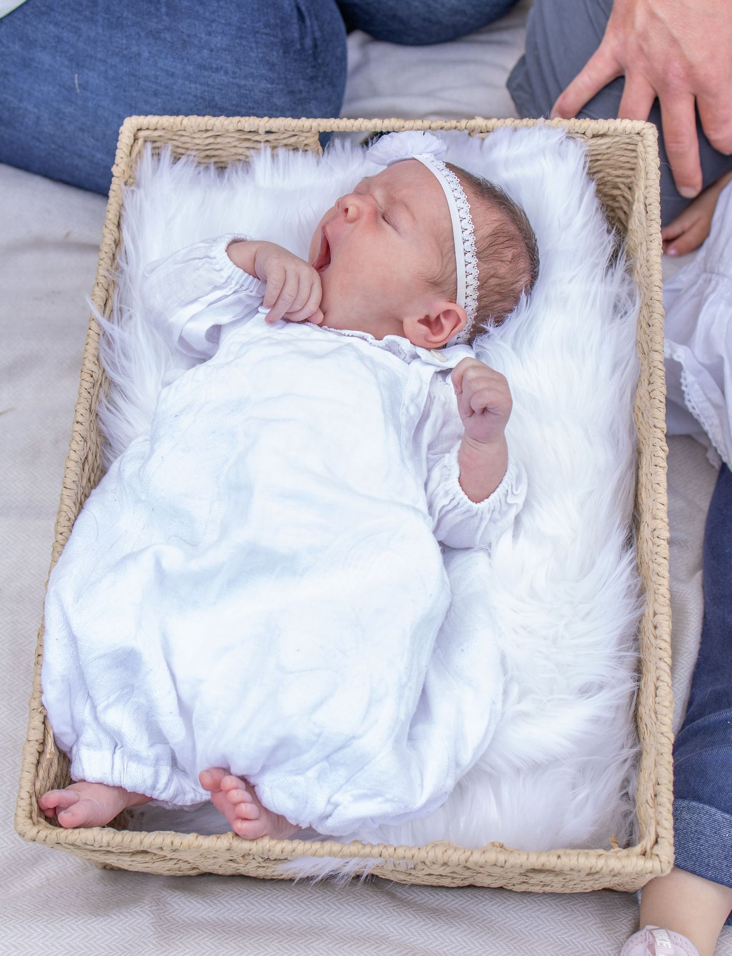 Newborn baby in white outfit and headband lying on fluffy white fur inside a wicker basket.