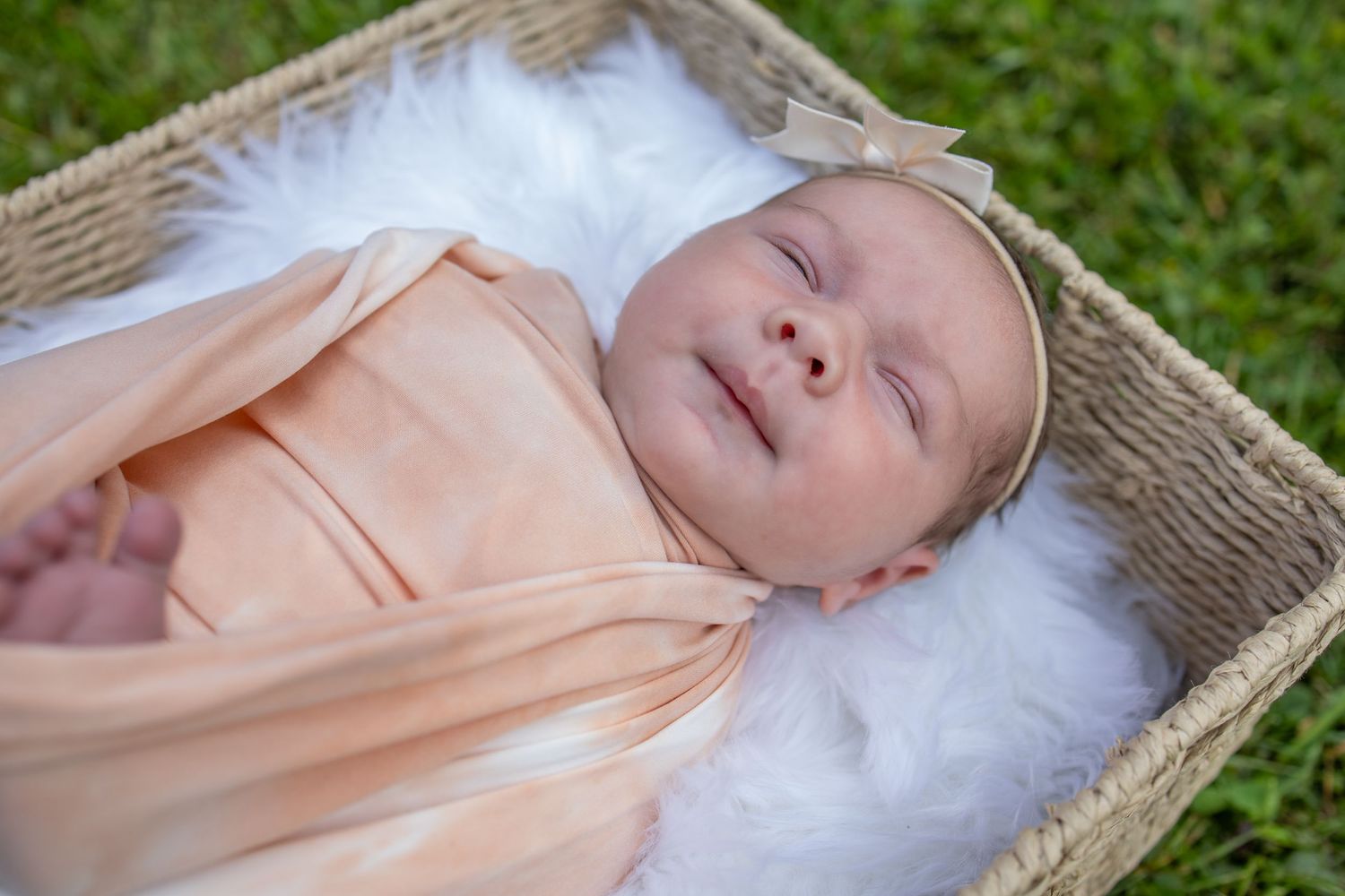 Newborn baby swaddled in peach wrap sleeping peacefully in a wicker basket lined with white fur outdoors.