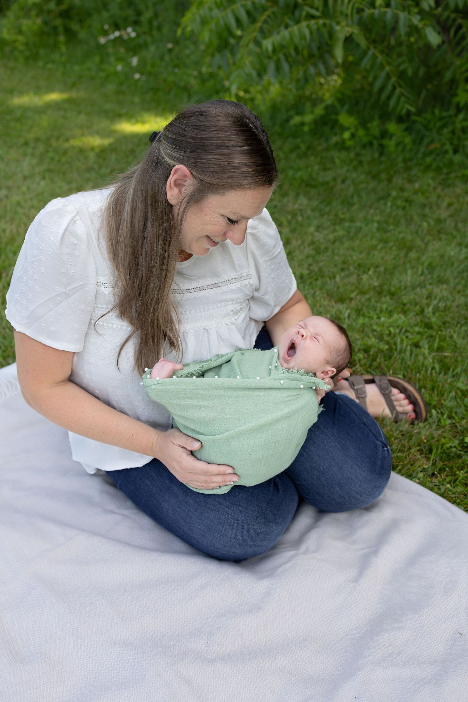A smiling mother sits on a blanket outdoors holding her yawning newborn baby wrapped in a green swaddle blanket.