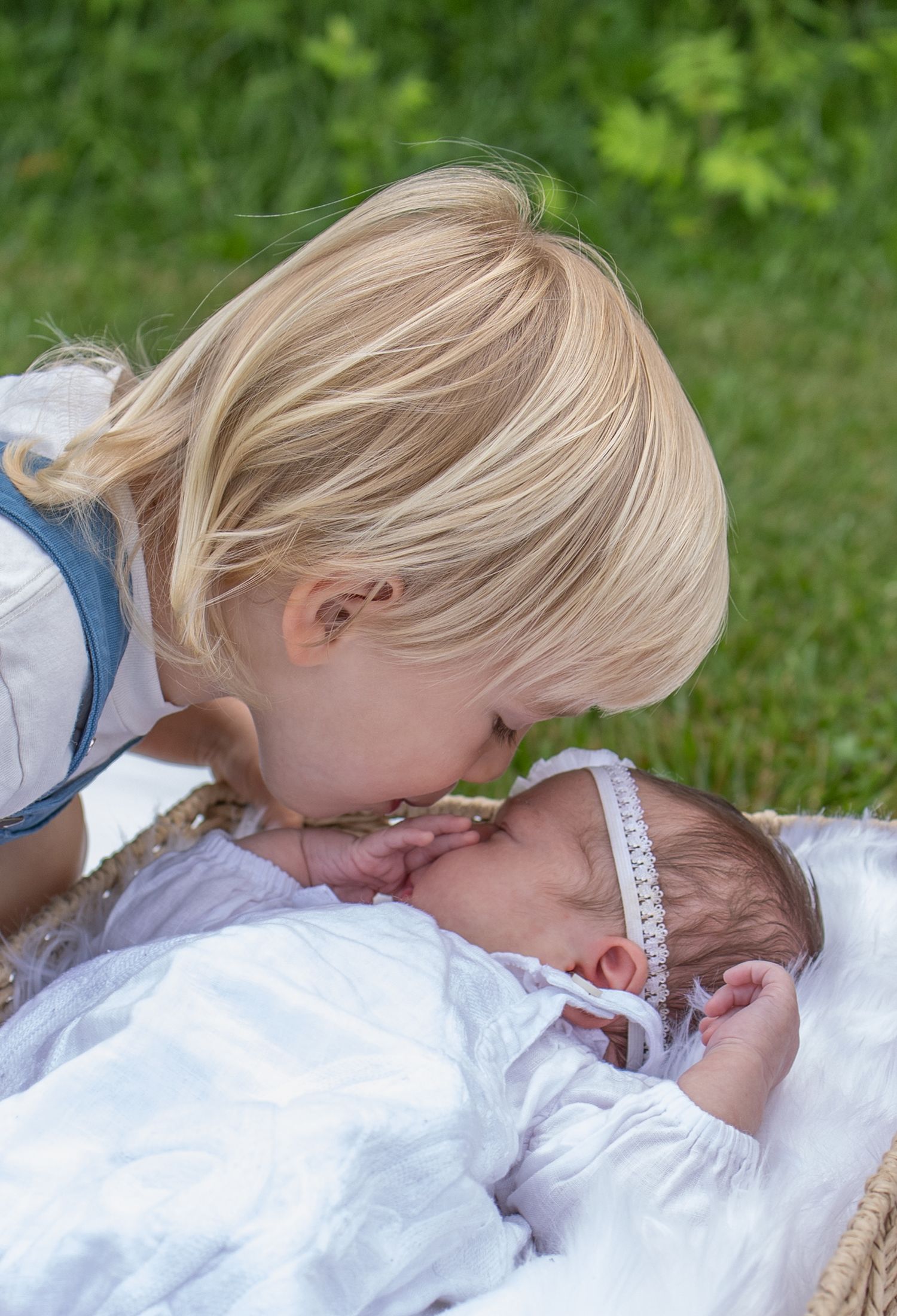 Blonde toddler gently kissing a sleeping newborn baby wearing a white headband, outdoors on green grass.