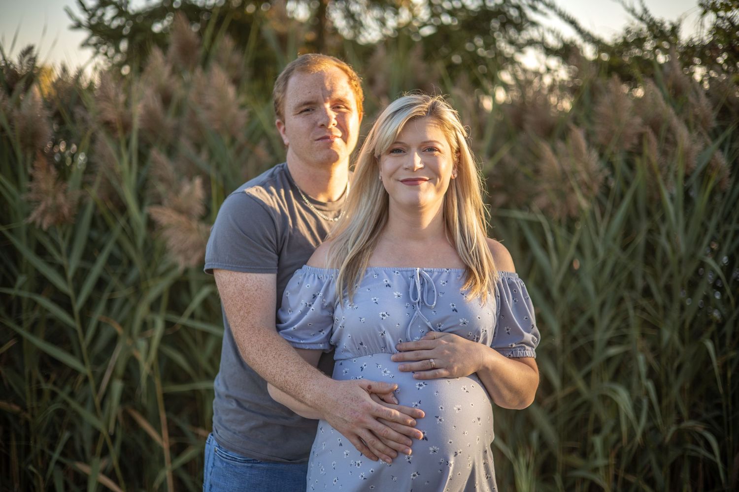 A couple embraces in front of tall grass during golden hour. During maternity session