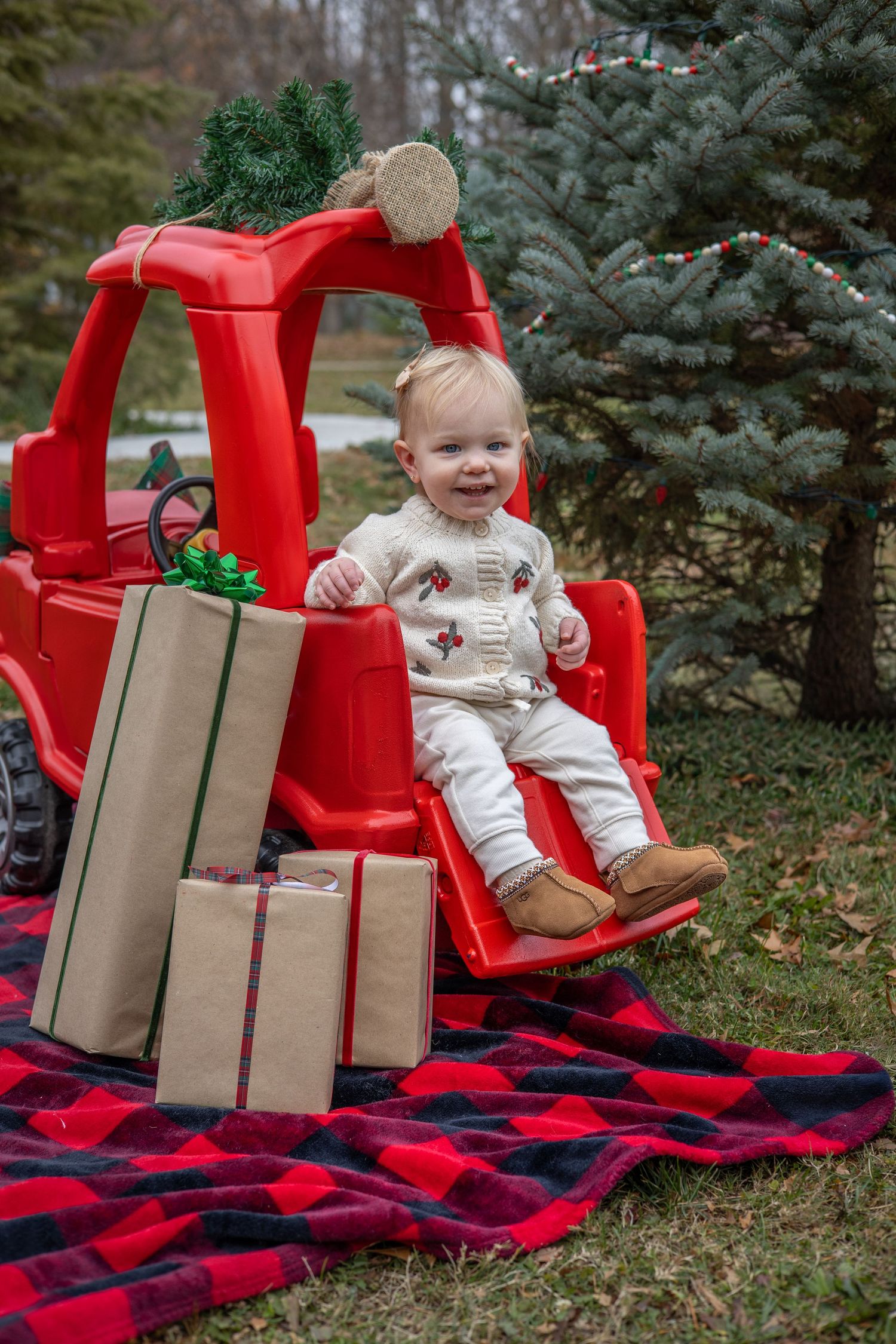 Holiday photo series with red truck, plaid blanket, and brown paper wrapped presents against evergreen trees. During Christmas Minis