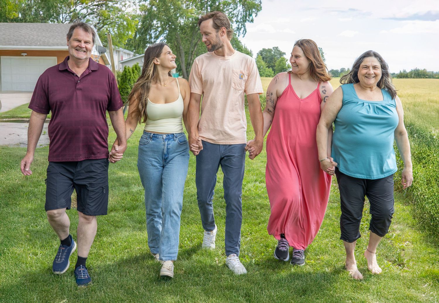 Five people walking together on green grass outdoors, smiling and holding hands on a sunny day.