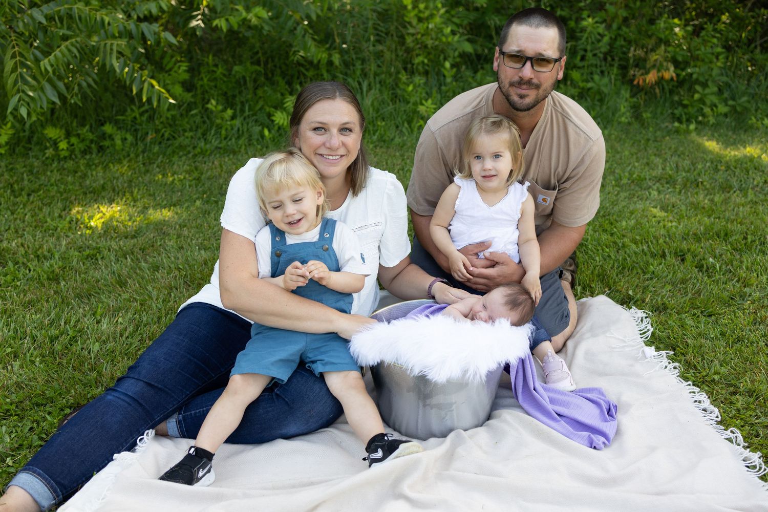 Family of four poses outdoors on blanket with newborn in bucket, two toddlers, lush green trees background.