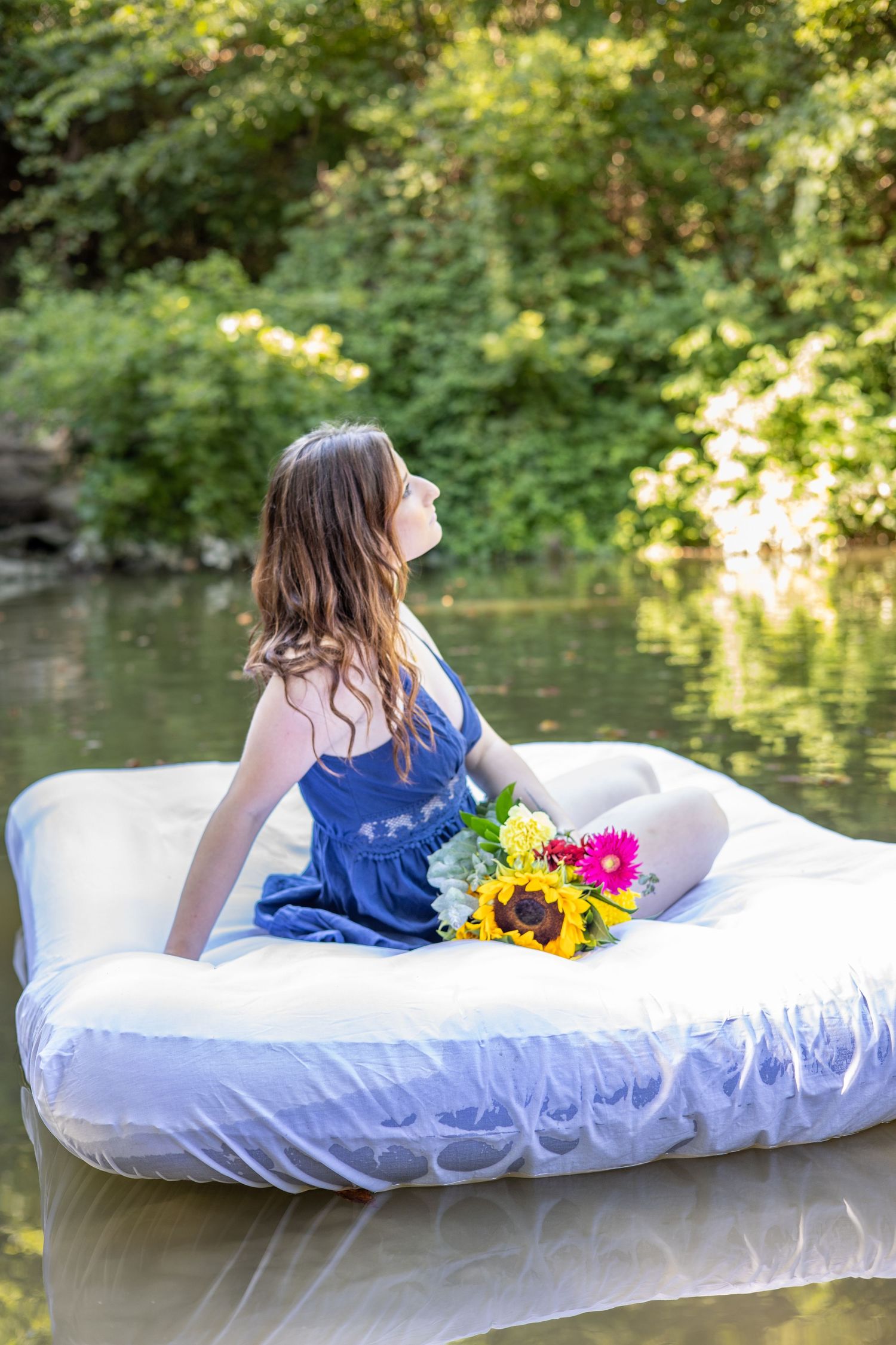 Person lounging on white mattress near pond with colorful flowers.