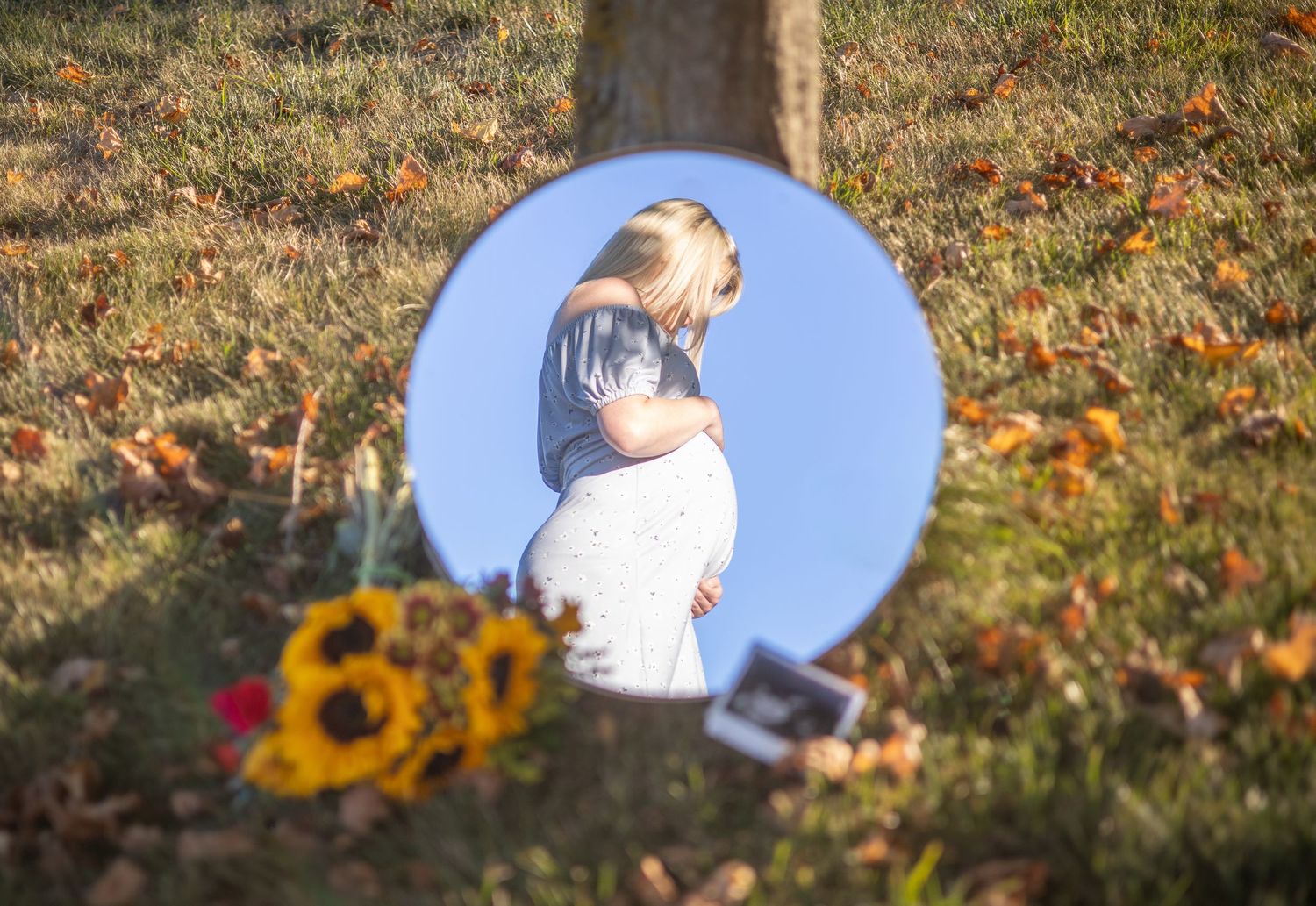A circular mirror reflects autumn leaves and sunflowers in an outdoor setting. During maternity session