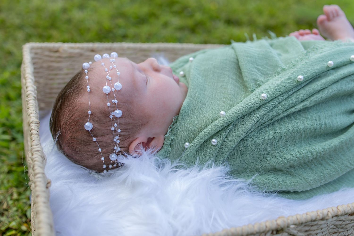 Newborn baby swaddled in green muslin wrap with pearl headband, sleeping peacefully in a wicker basket.