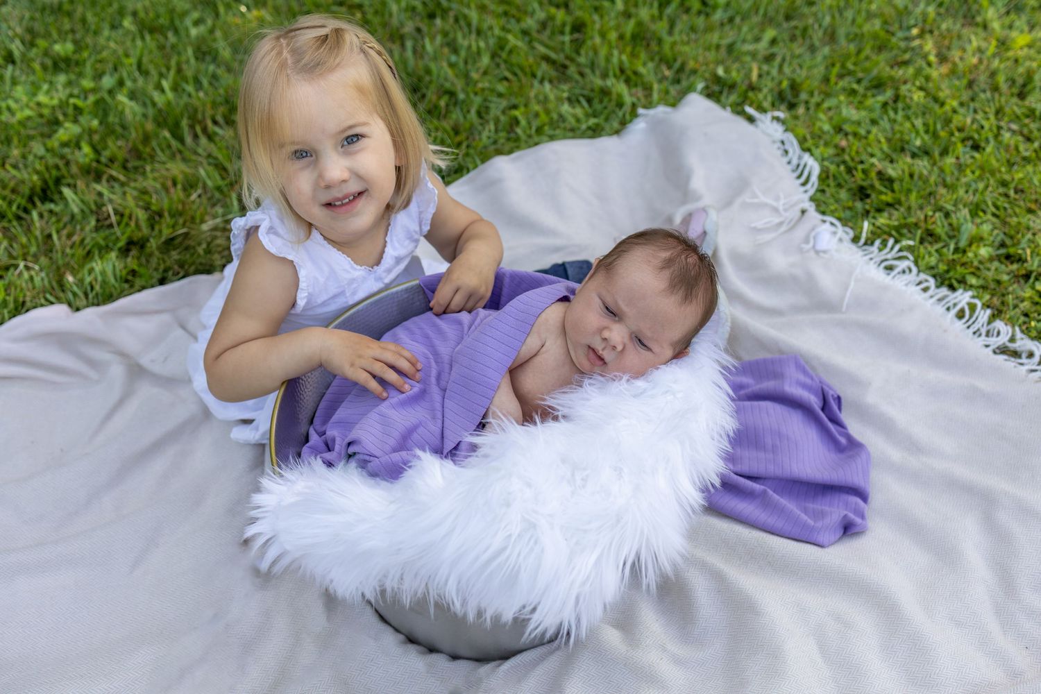 Young girl smiling beside newborn baby wrapped in purple swaddle in fluffy white basket on white blanket outdoors.