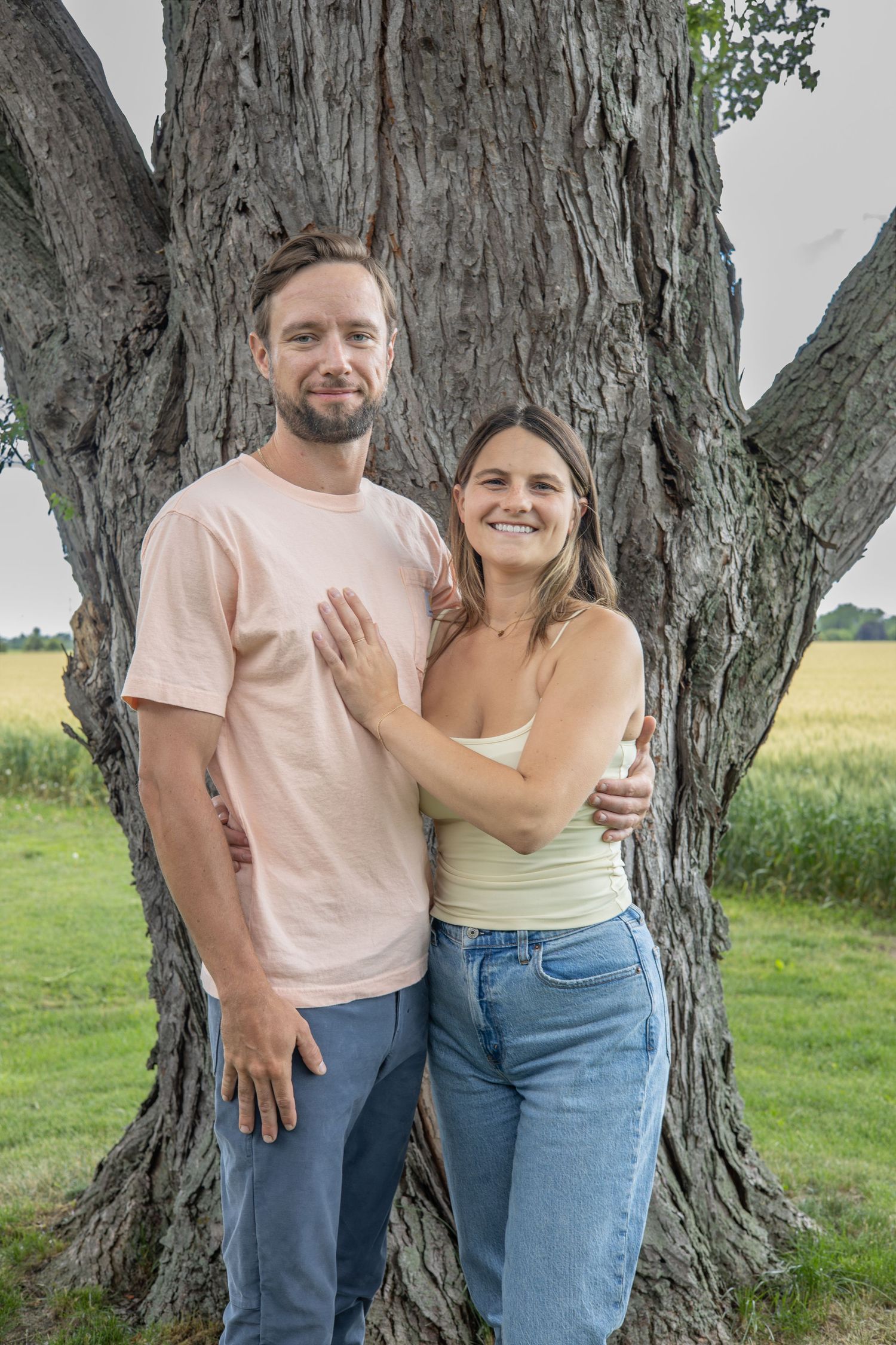 A smiling couple poses together in front of a large tree with green fields in the background on a summer day.