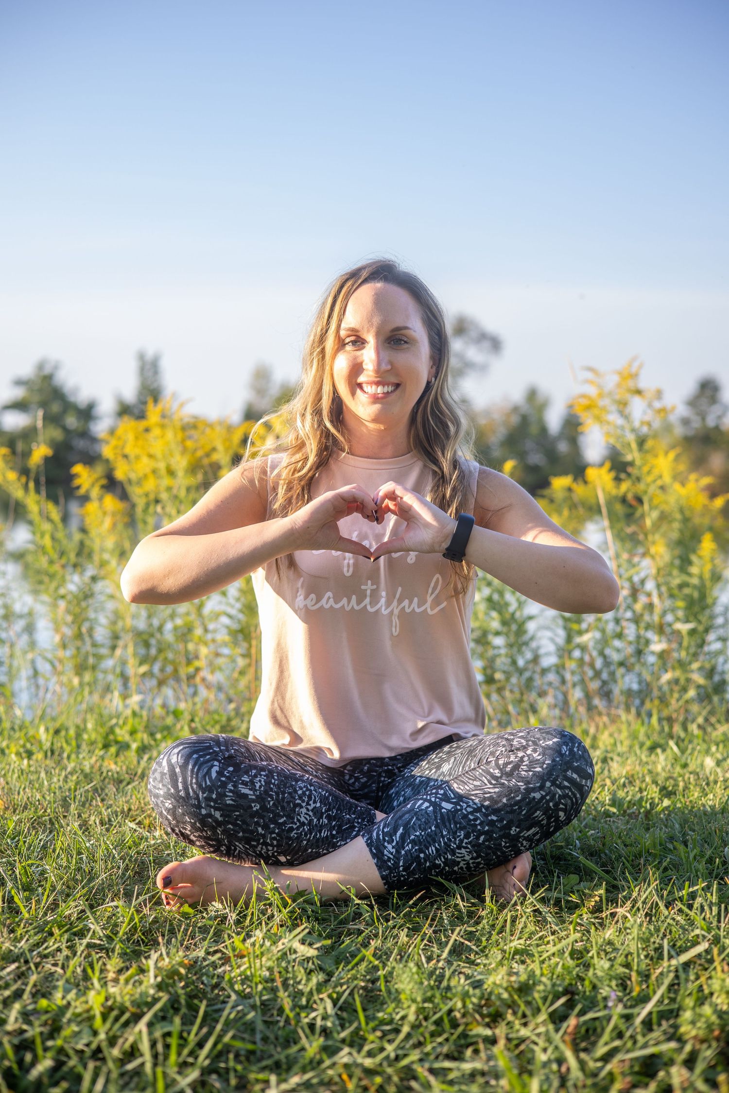 Woman sitting in field of yellow wildflowers on sunny day making heart shape with hands.