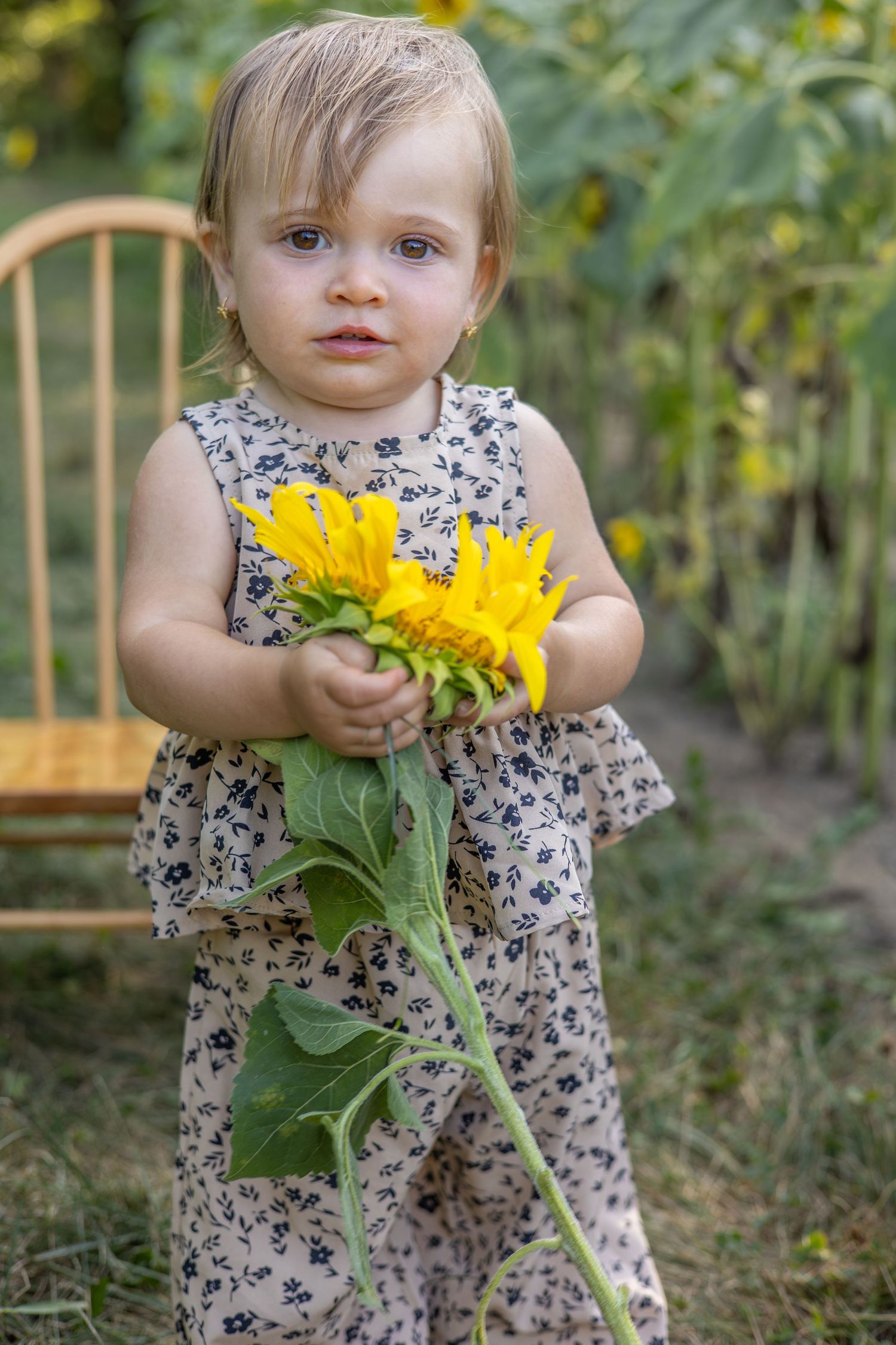 Toddler girl in floral dress holding a sunflower outdoors near a wooden chair.