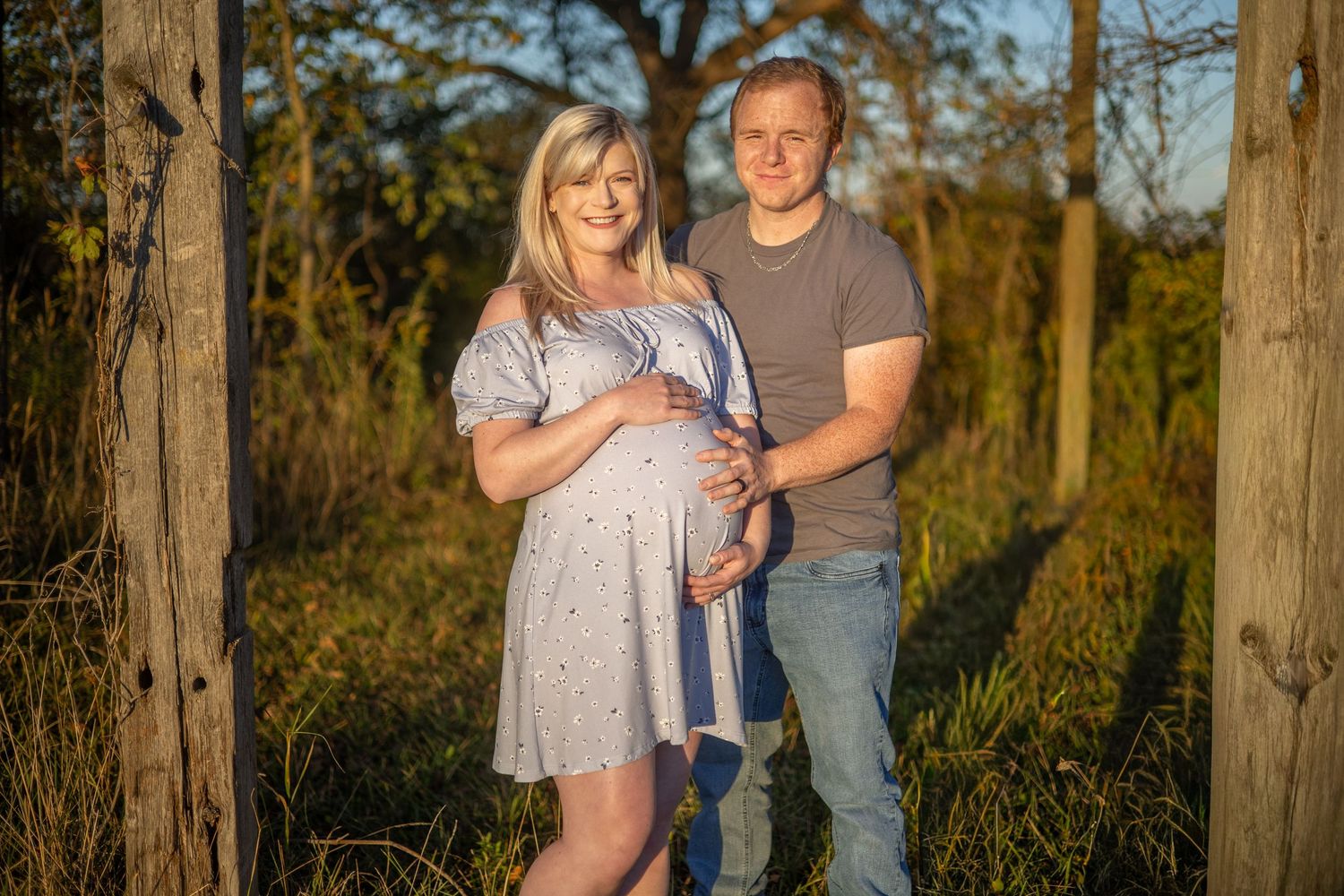 A couple poses together between trees in warm evening light.