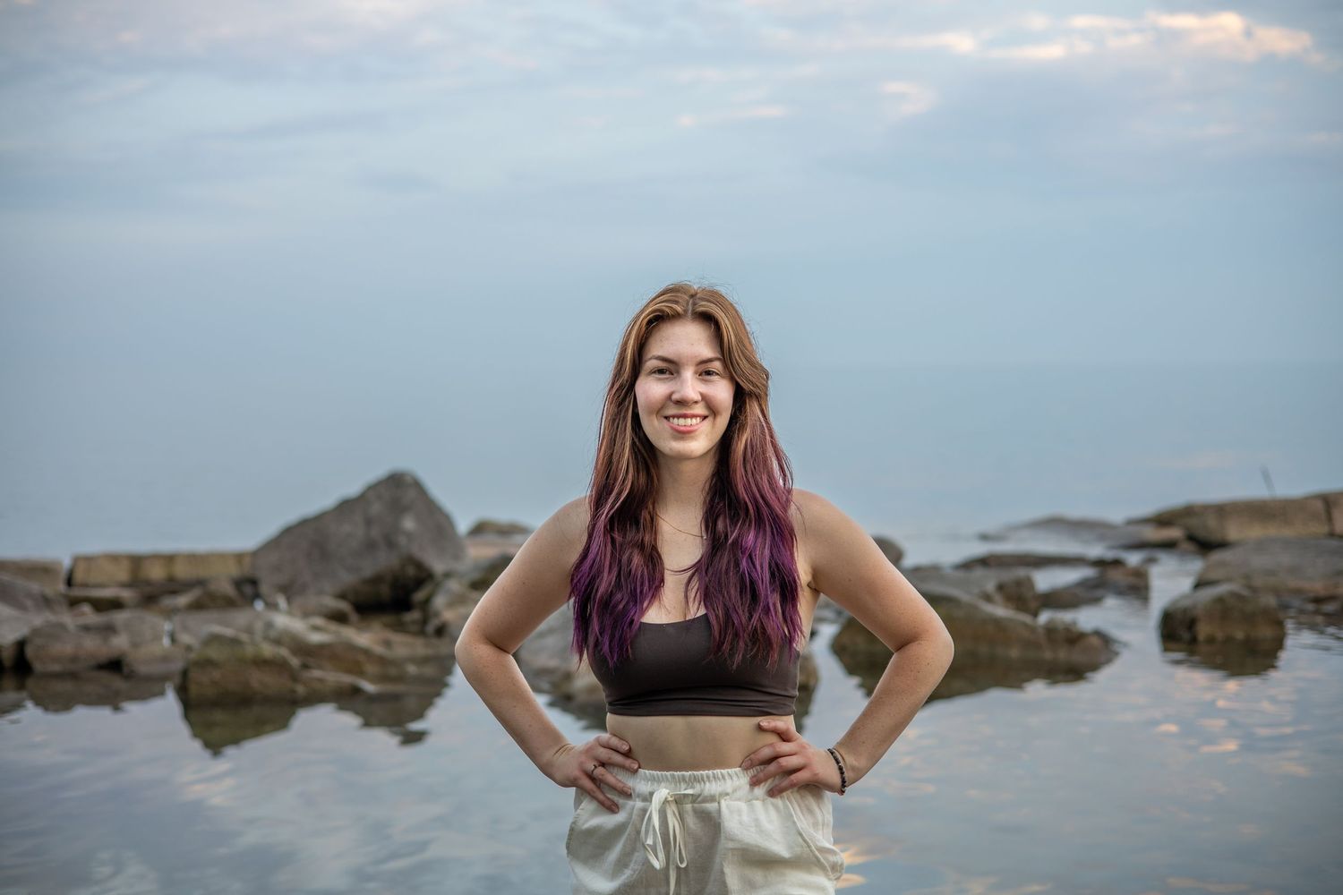Person posing on rocky shoreline with water and cloudy sky in background.