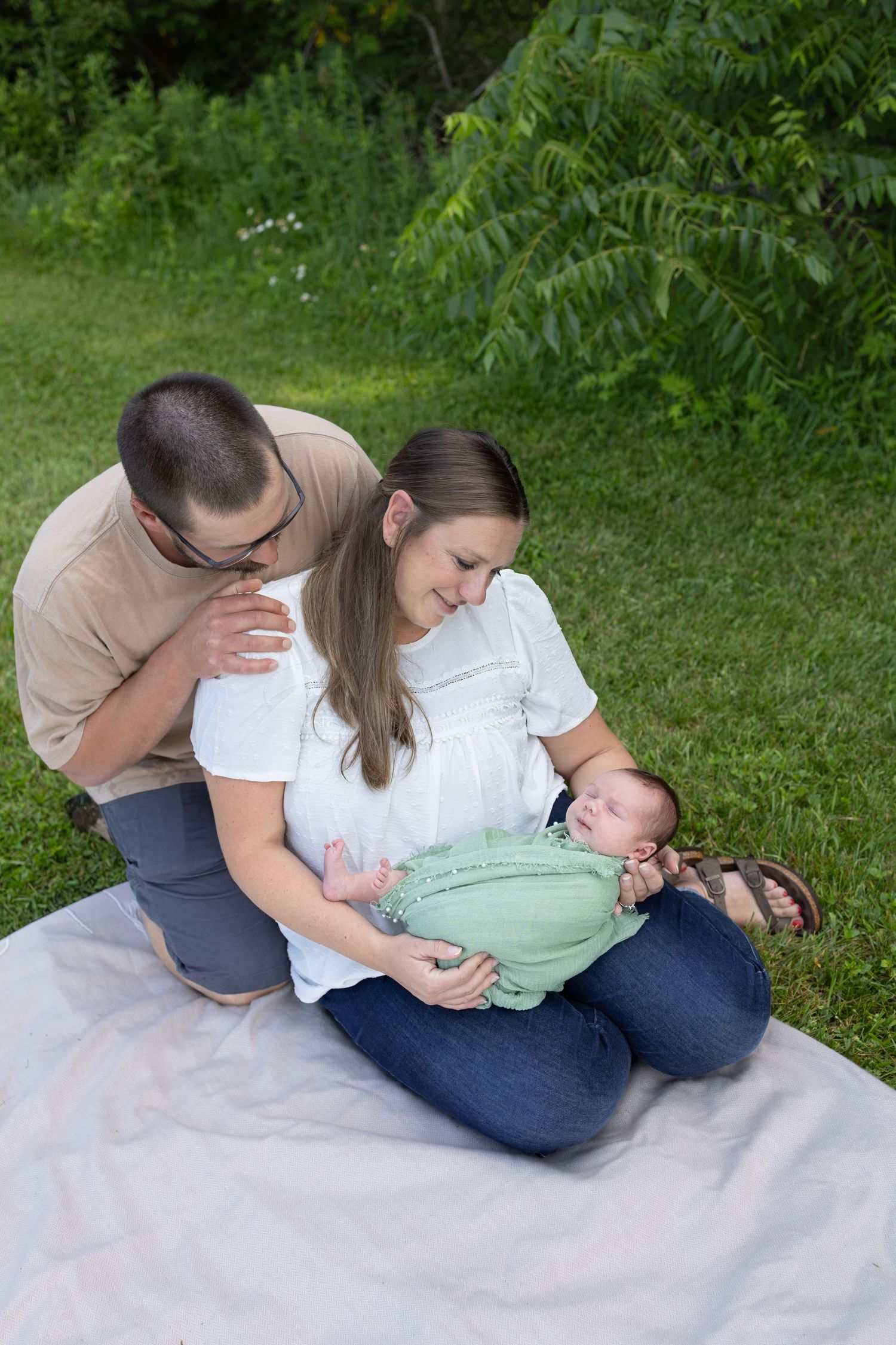 Parents sitting on a blanket outdoors admiring their newborn baby wrapped in a green swaddle blanket.