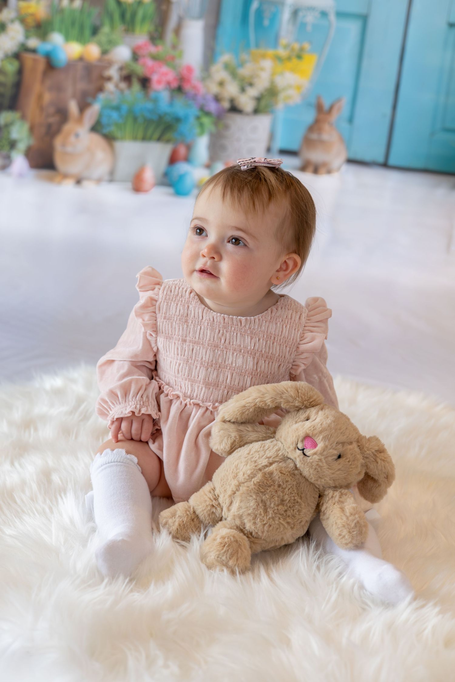 Baby girl in pink dress sits on white fur rug with bunny toy, surrounded by Easter decorations and live rabbits.