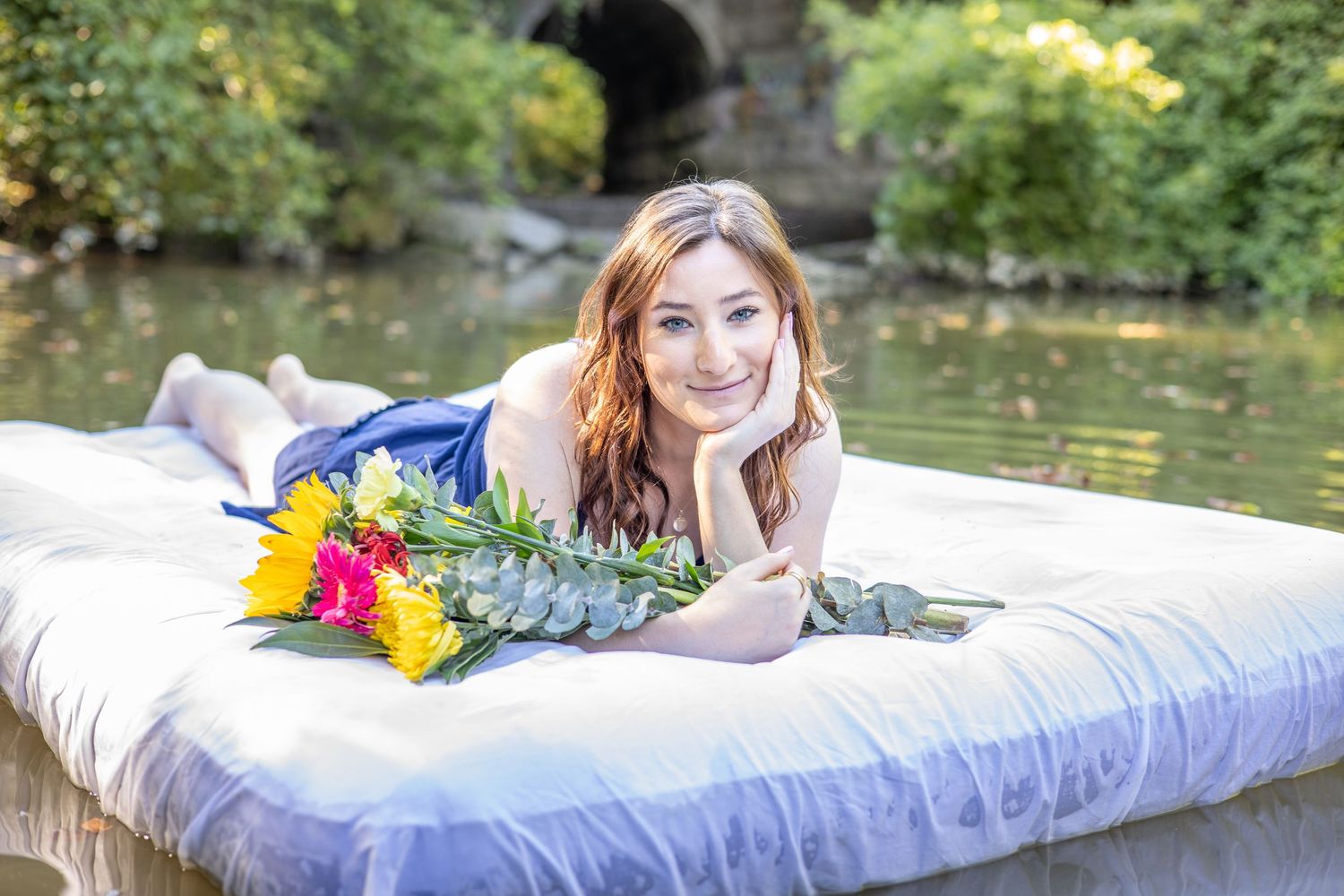 A mattress with floral decorations sits in a lush garden setting near a pond.