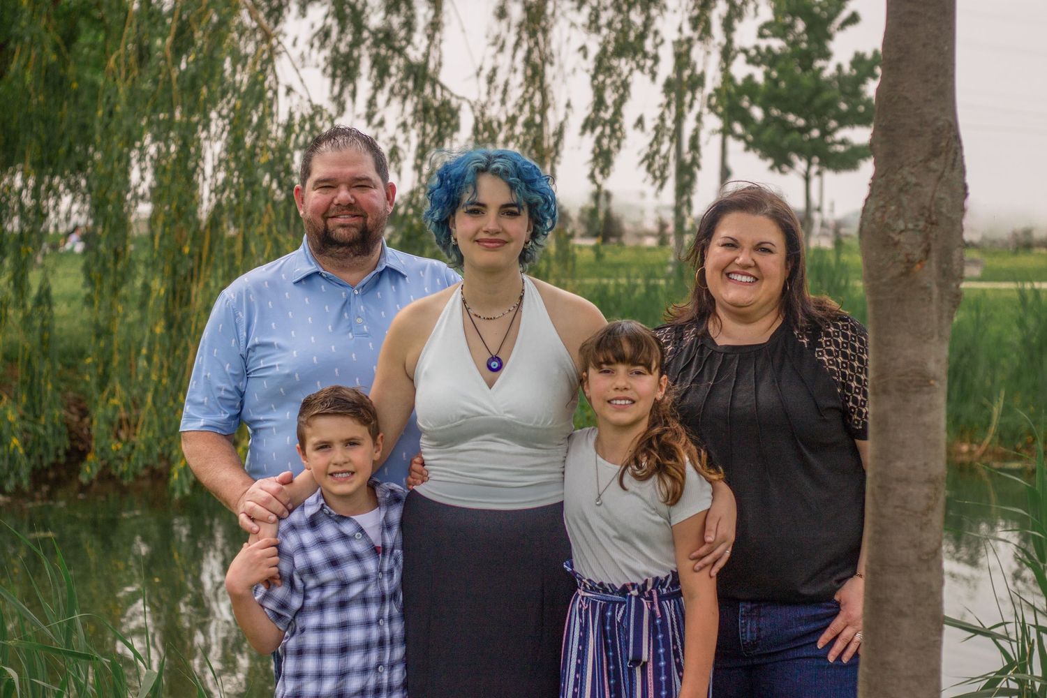 A family group poses together outdoors near trees and water on a sunny day.