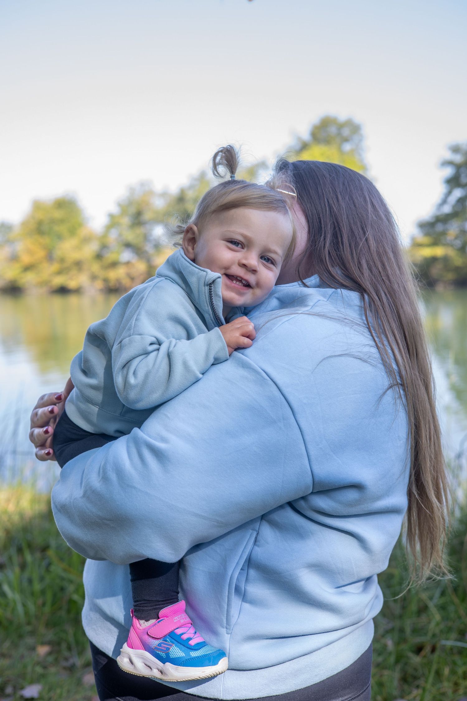 Two people in light blue hoodies embrace outdoors near a lake.