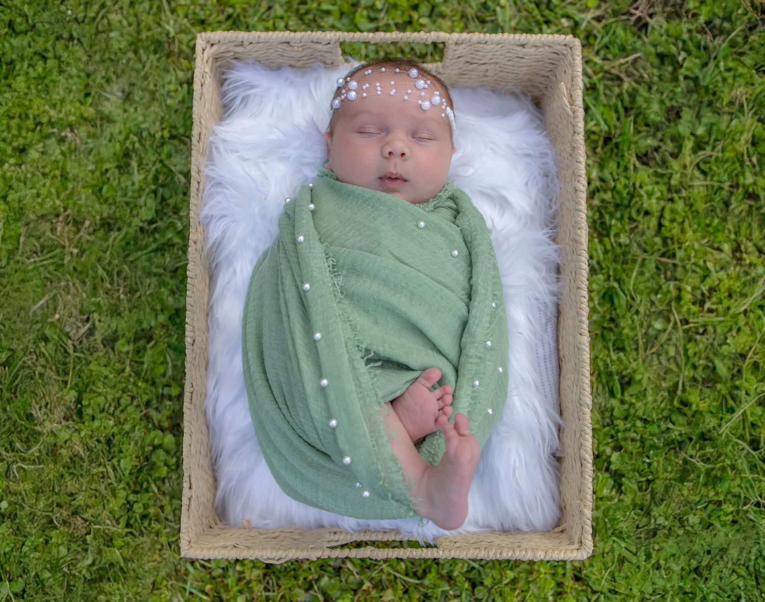 Newborn baby swaddled in green wrap with pearl accents, sleeping in a wooden box lined with white fur on grass.