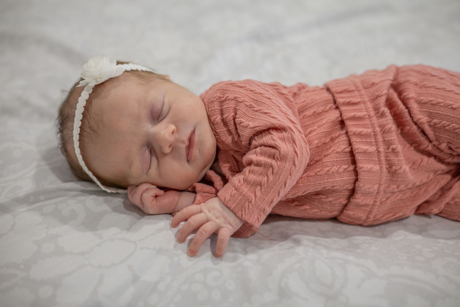 A sleeping baby wearing a peach-colored knit outfit and white headband lies on soft white bedding.