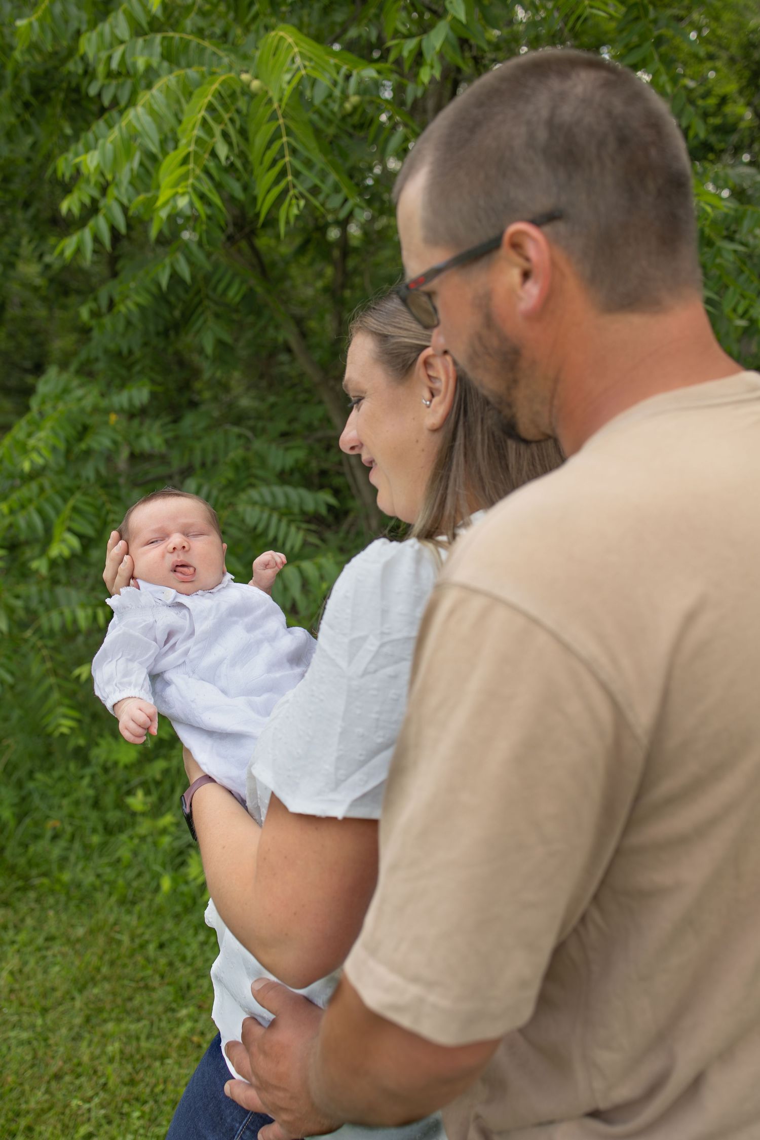 Happy parents holding their newborn baby outdoors surrounded by lush green trees.