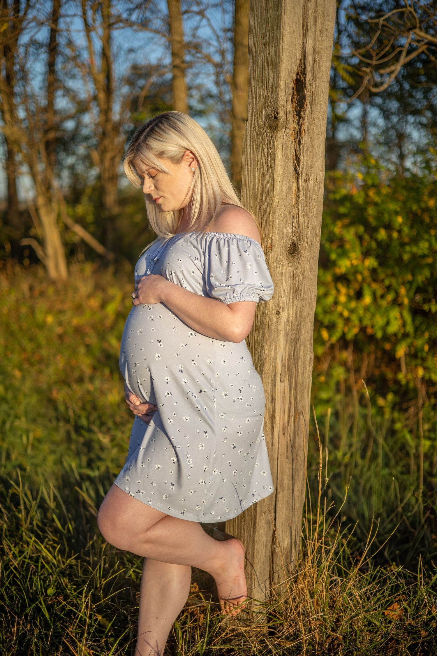A pregnant woman in a light dress leans against a tree trunk in golden sunlight.