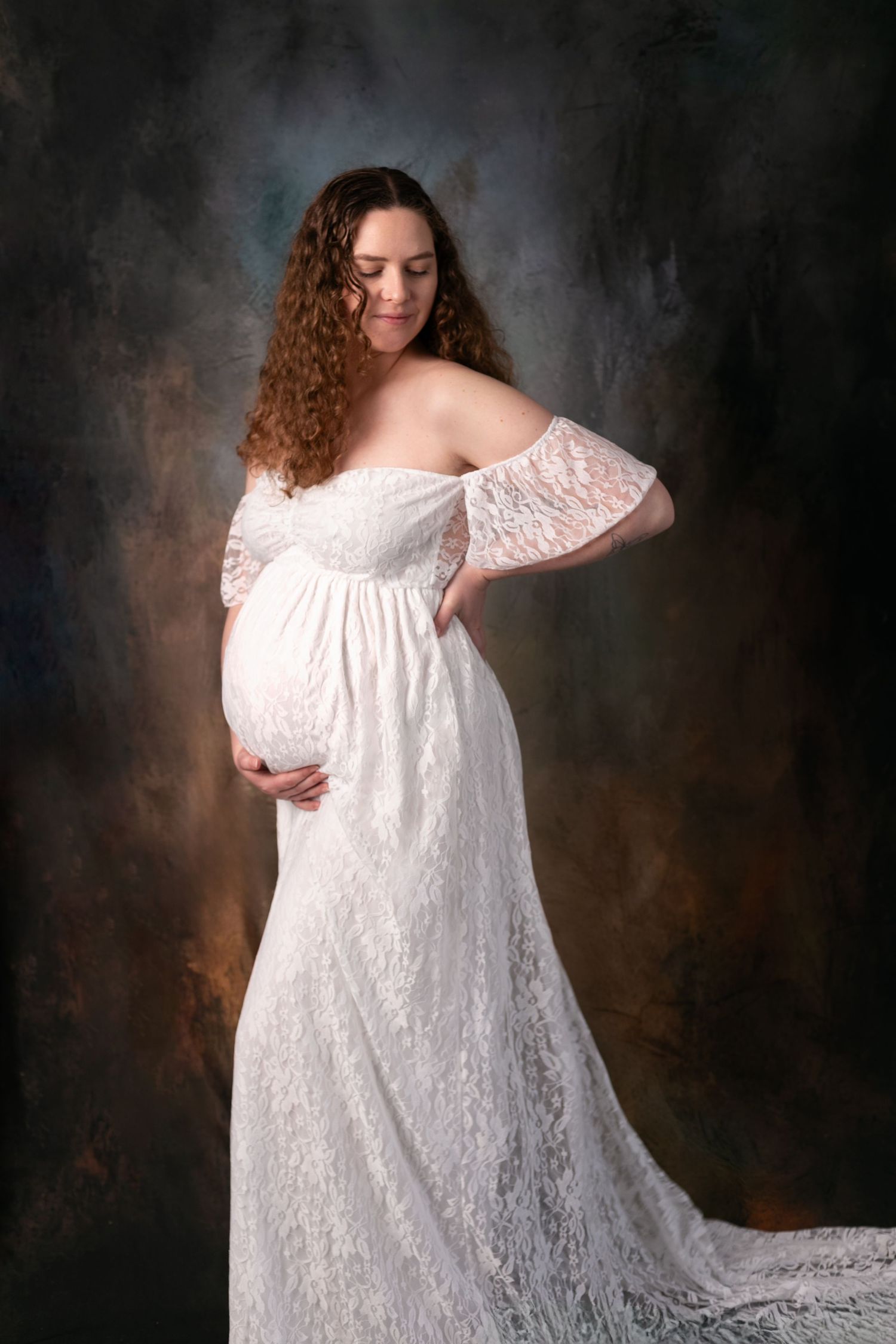 A maternity portrait series showing a flowing white lace dress against a dark dramatic studio backdrop.