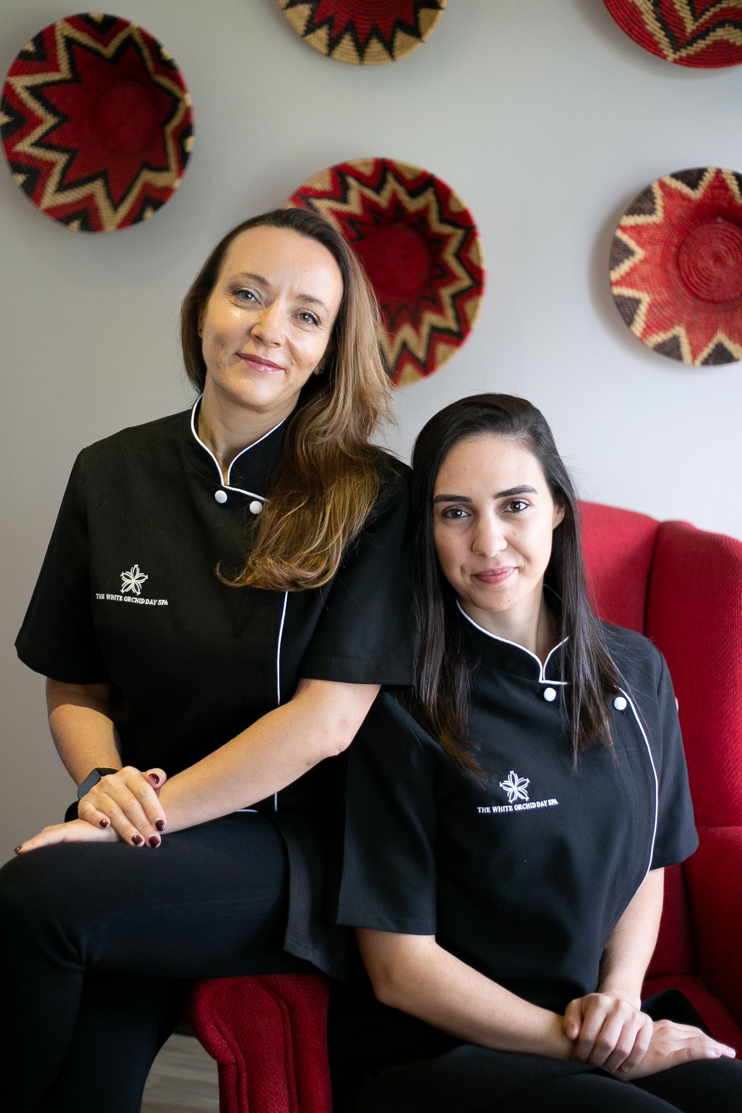 Two women in black spa uniforms pose together in front of decorative woven wall art in red and natural tones.