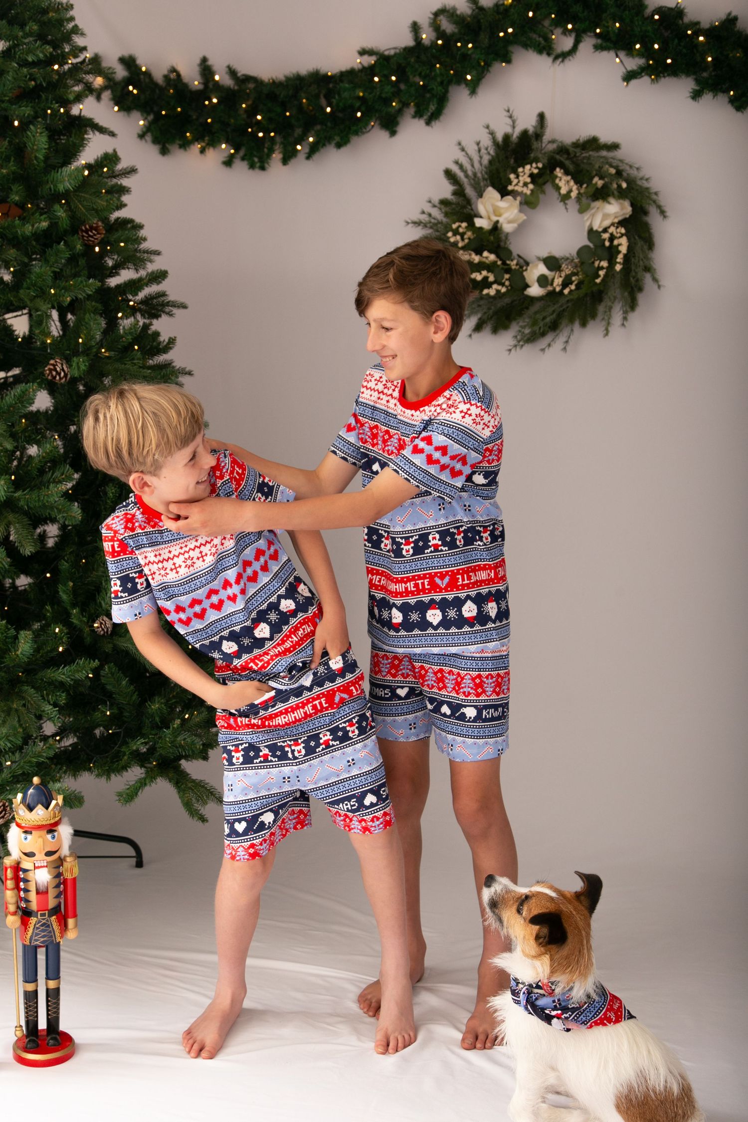 Children in matching holiday pajamas play with a decorative nutcracker by a Christmas tree in a white room.