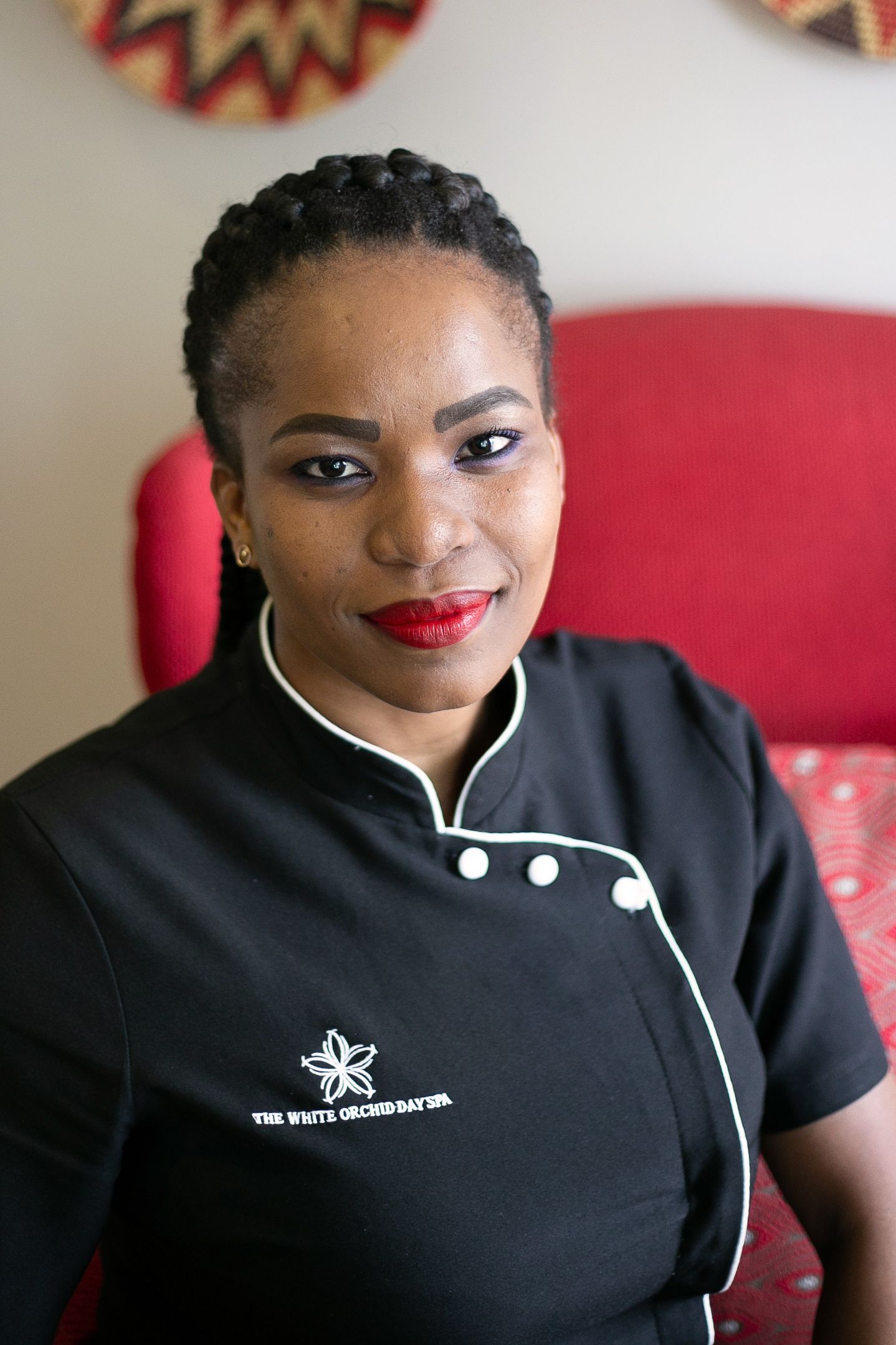 Woman in black White Orchid Day Spa uniform with braided hair and red lips, seated in front of red chair.