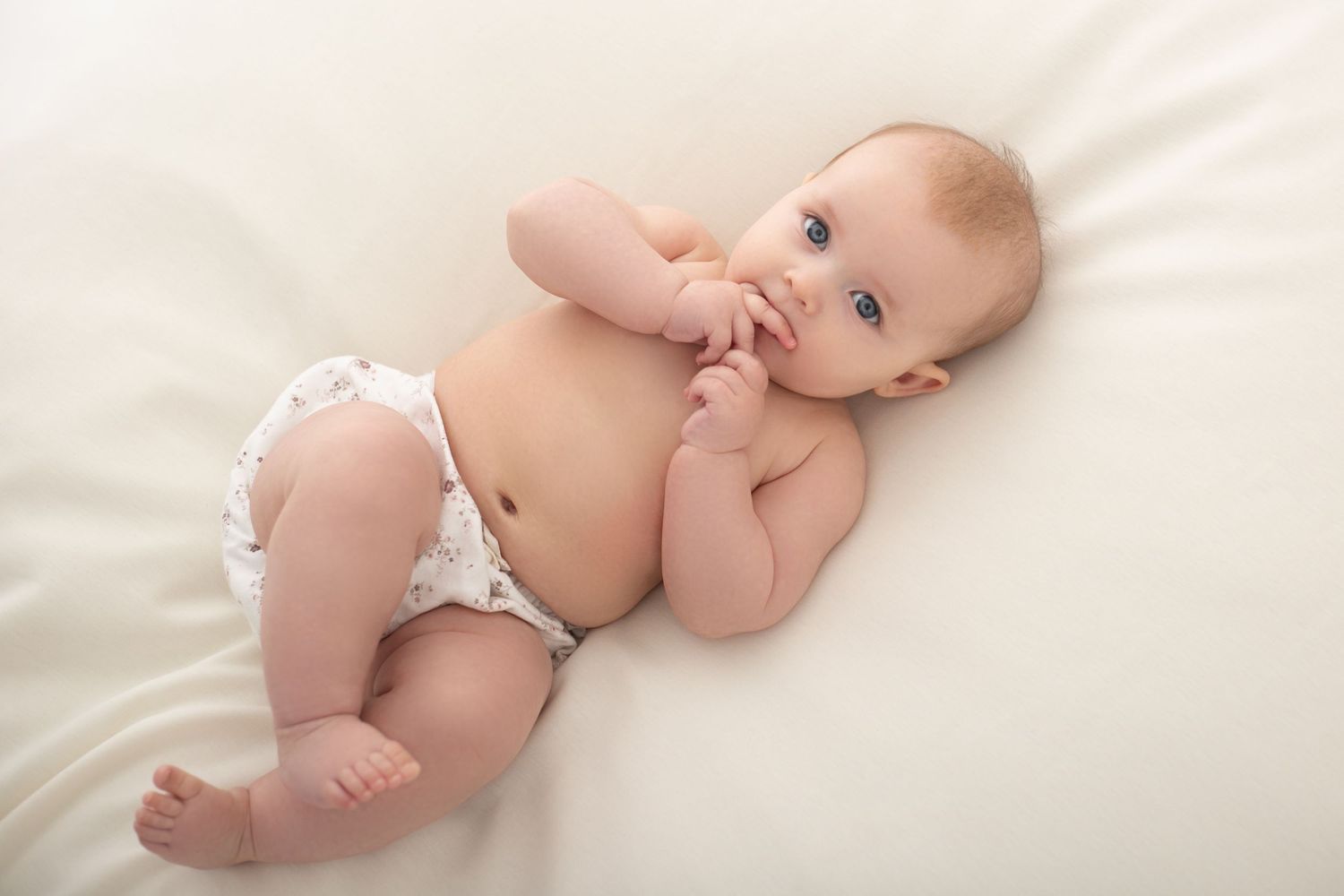 Cute baby lying on white bed wearing a diaper, looking up with blue eyes and fingers in mouth.