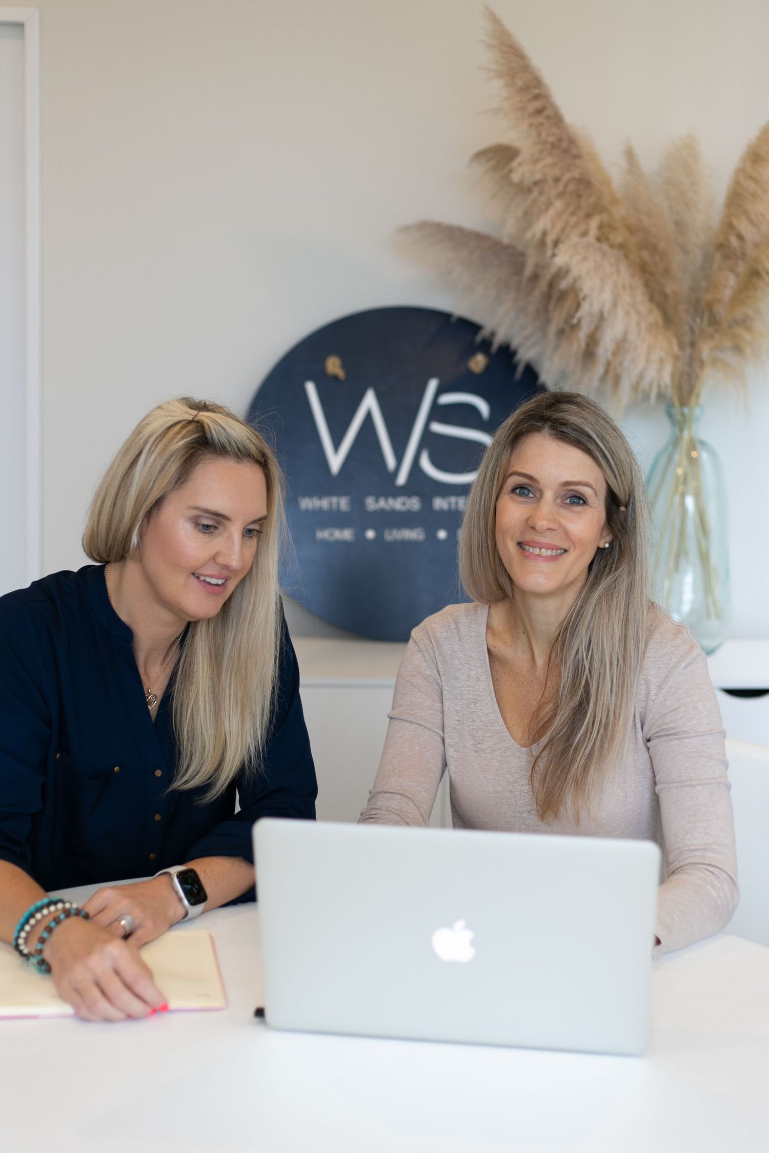 Two women collaborating on a MacBook laptop at White Sands Interiors office with pampas grass decor.