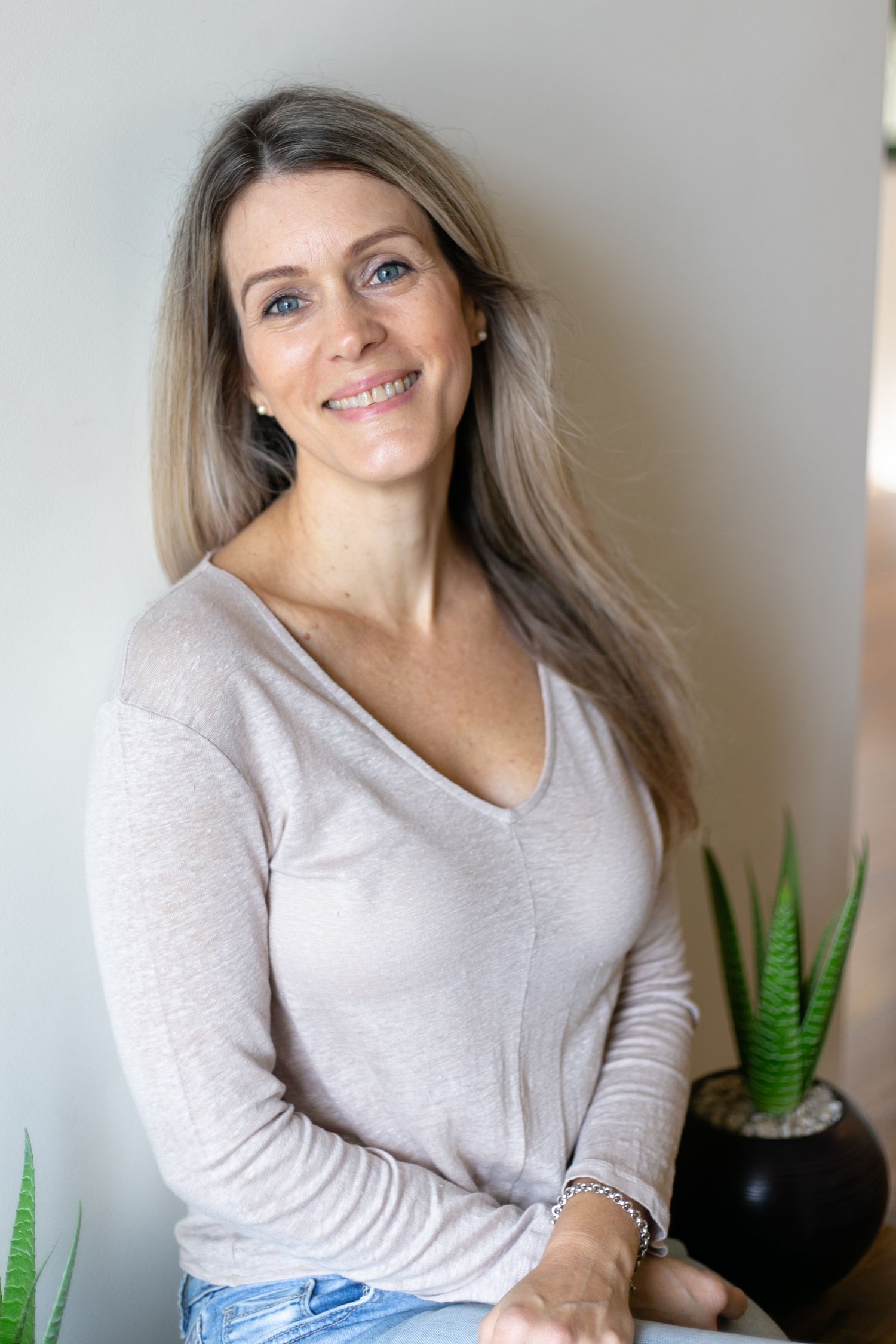 Smiling woman with long blonde hair wearing a beige v-neck sweater and jeans, posing indoors near a plant.