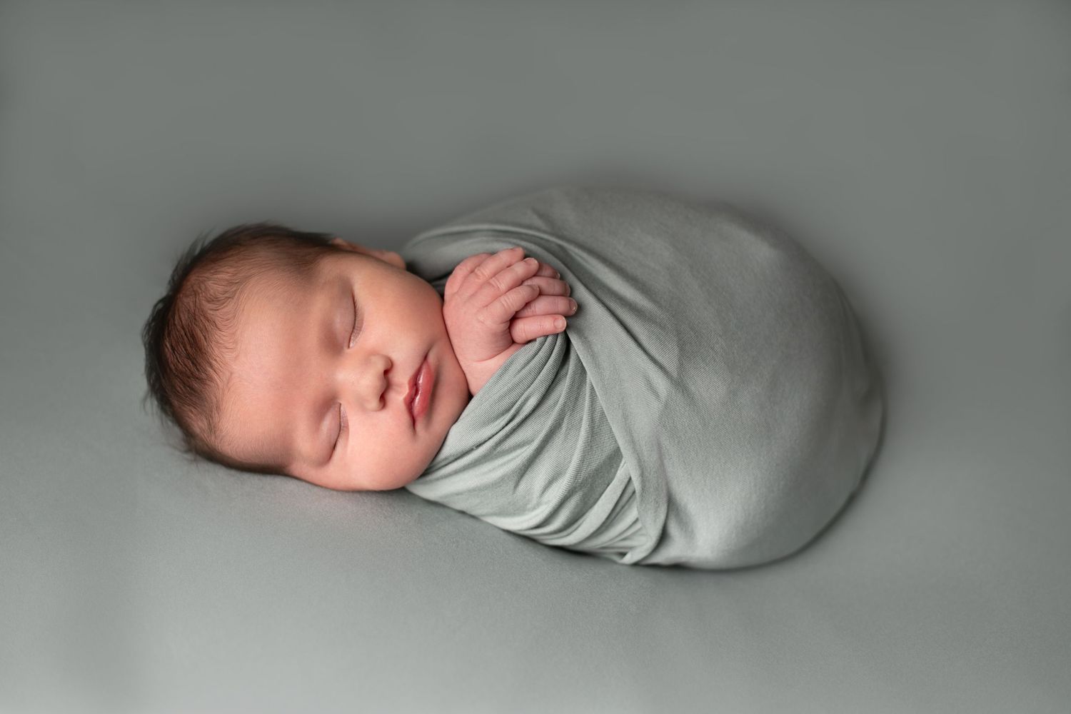 A sleeping newborn baby wrapped in a soft gray swaddle blanket lies peacefully on a gray backdrop.