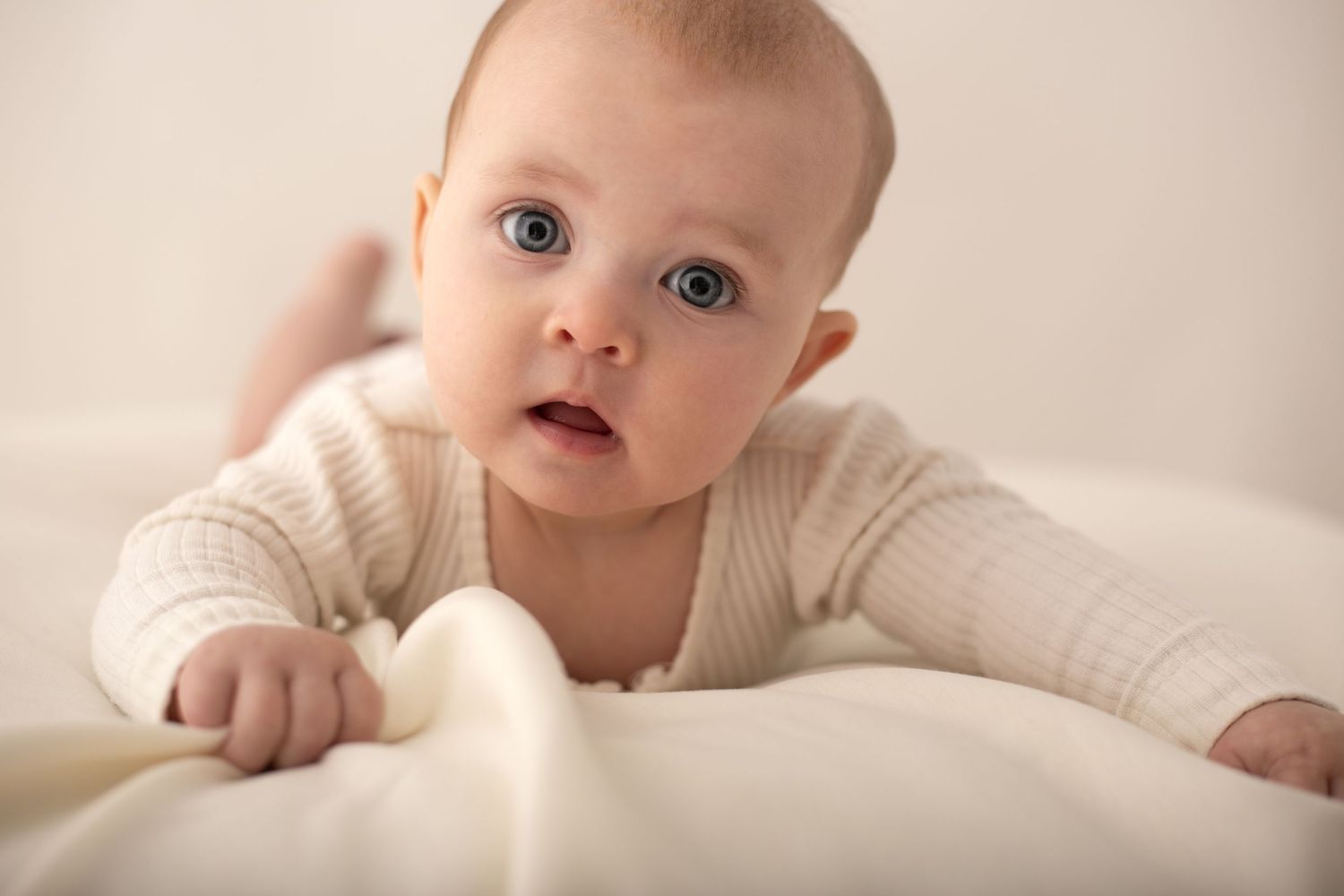 Adorable blue-eyed baby in a cream ribbed onesie doing tummy time on a soft white blanket.