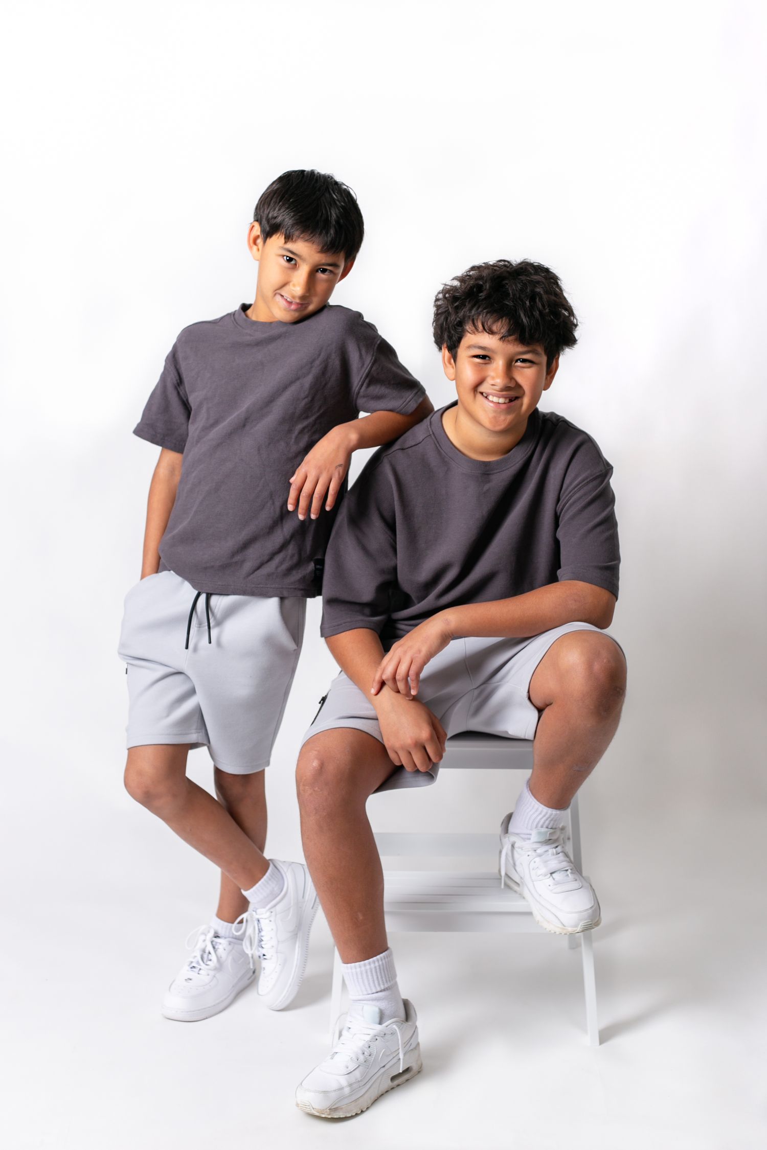Two people in matching gray shirts and white shorts sit on chairs in studio setting.