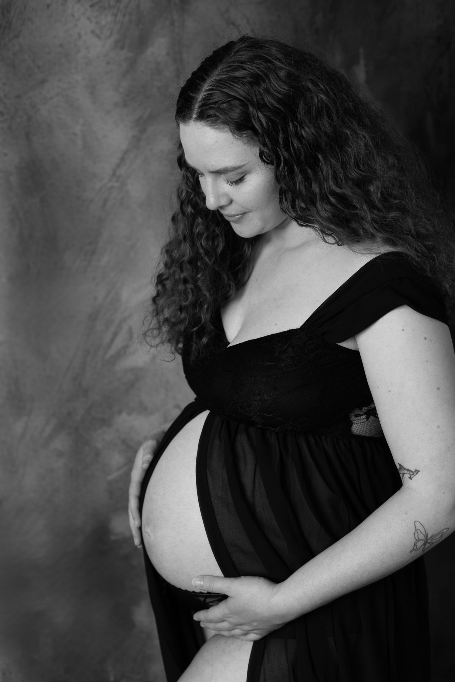Black and white artistic maternity photo series showing woman in dark dress cradling pregnant belly against gray backdrop.