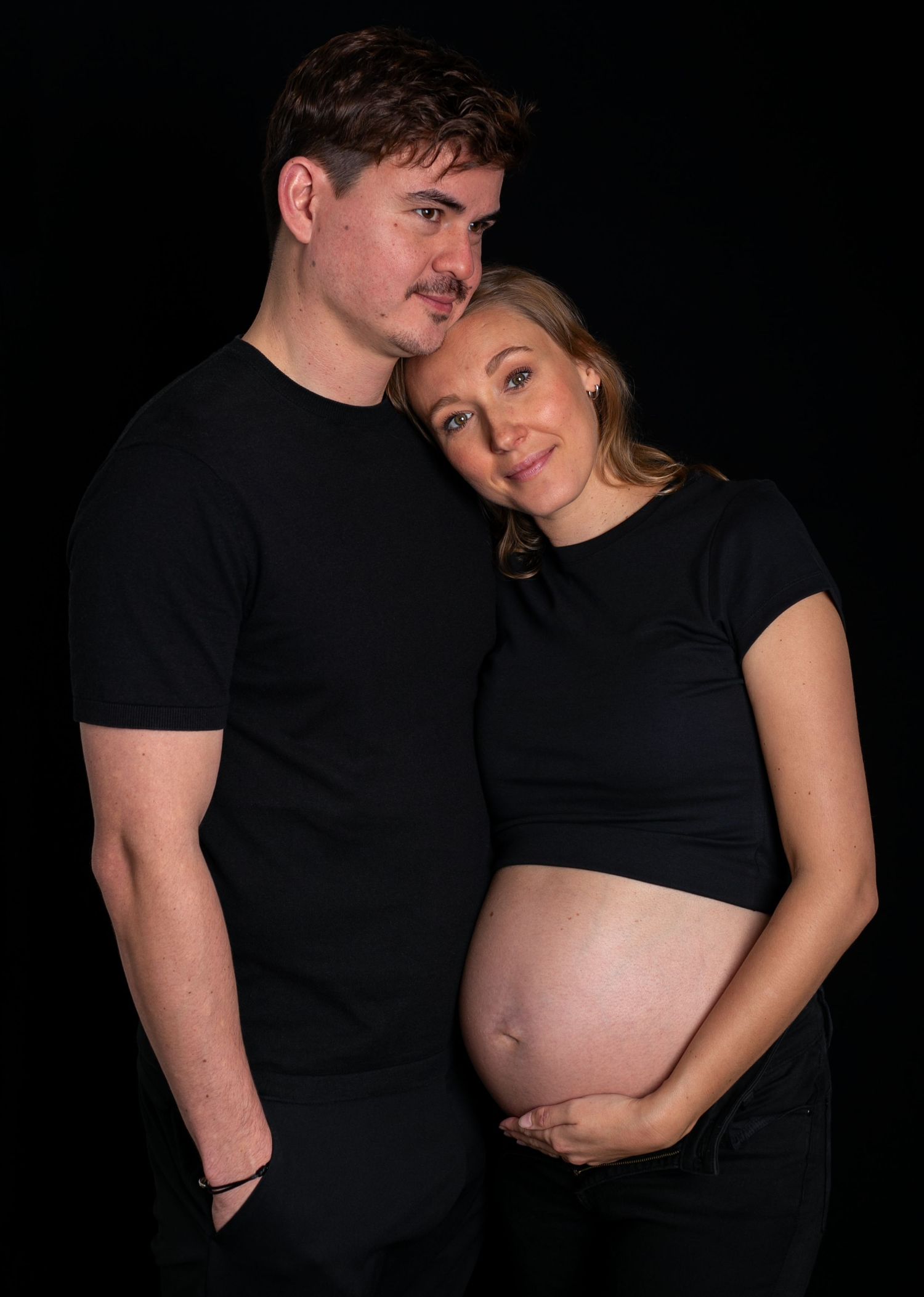 A couple in black clothing poses for a maternity photo series showing a growing baby bump against a dark background.