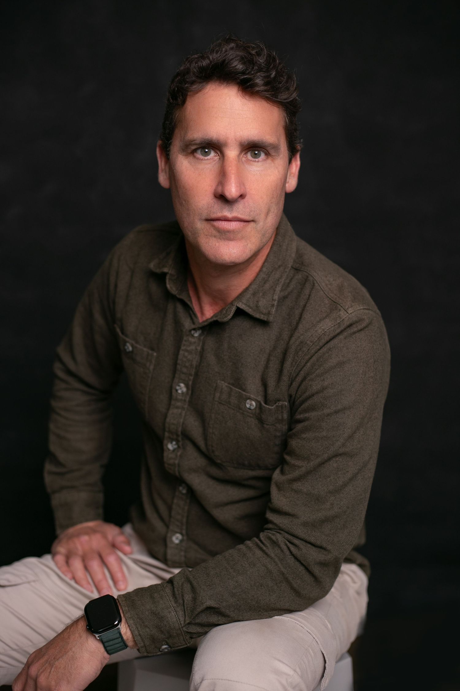 Man in olive green shirt and smartwatch sits confidently against dark studio background for professional portrait.