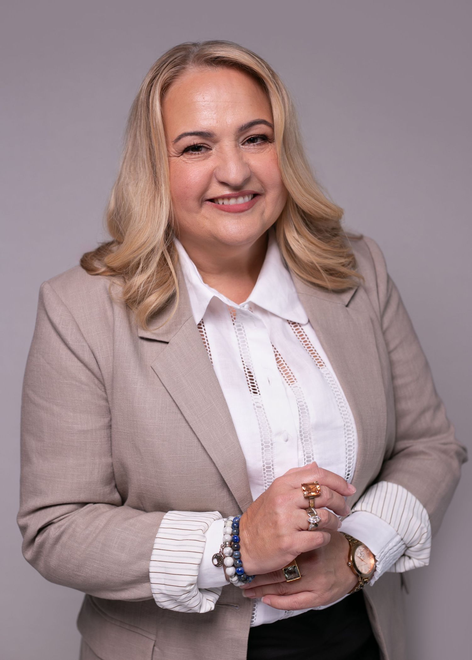 Professional woman in beige blazer and white blouse smiling confidently, wearing jewelry, against gray background.