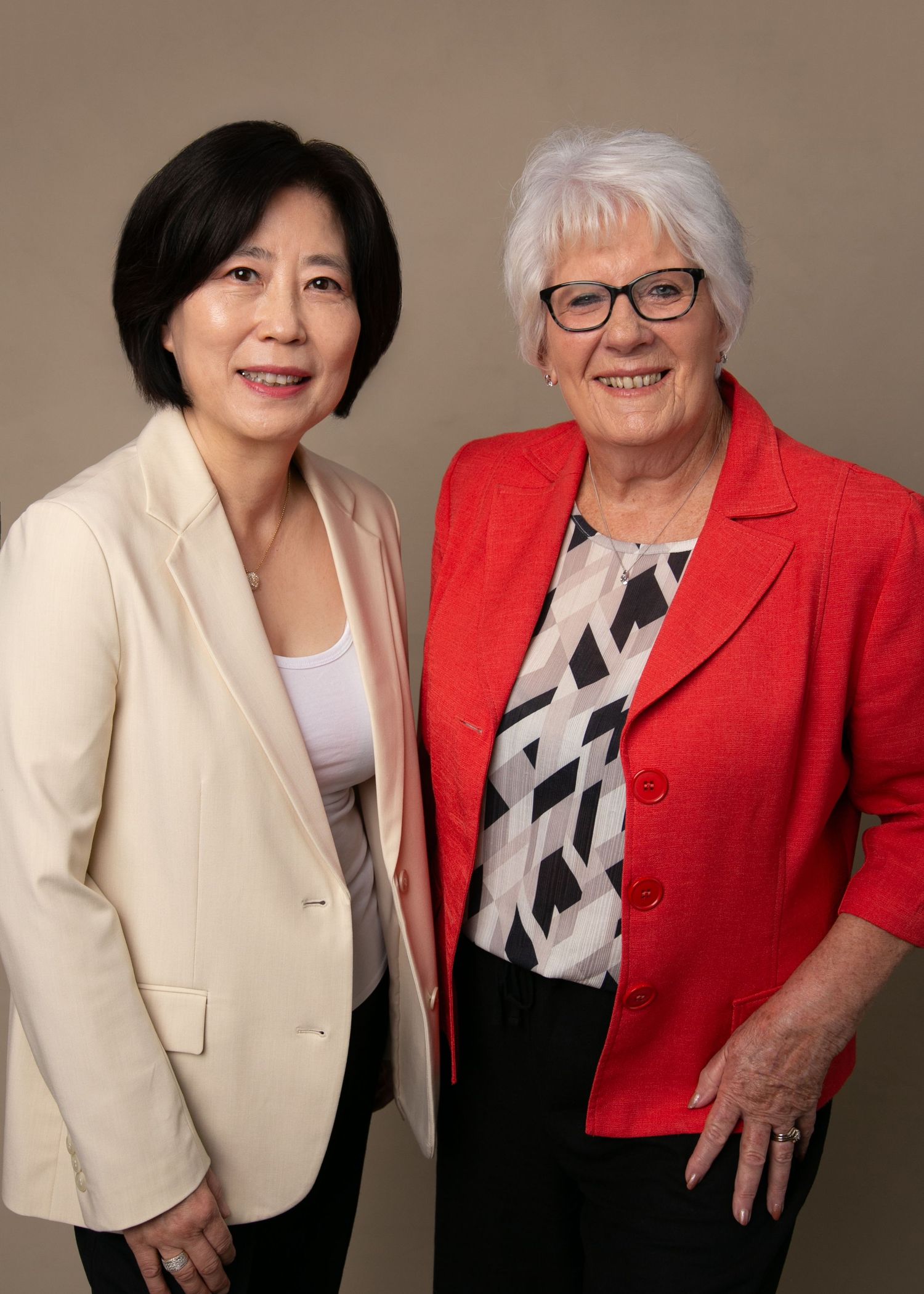 Two professional women smiling, one in a cream blazer and one in a red jacket, posing together for a formal portrait.
