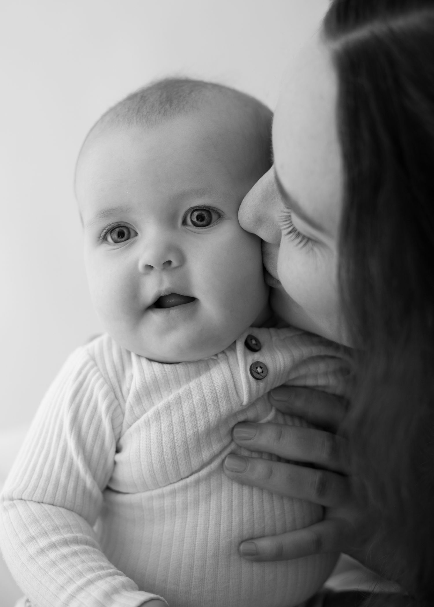 Black and white photo of a mother kissing her smiling baby on the cheek while holding them close.