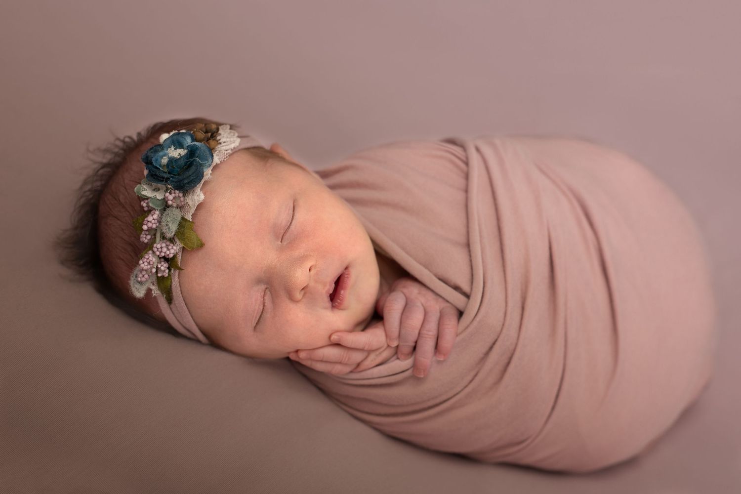 Peaceful sleeping newborn swaddled in soft pink wrap wearing delicate floral headband with teal accent on mauve backdrop.