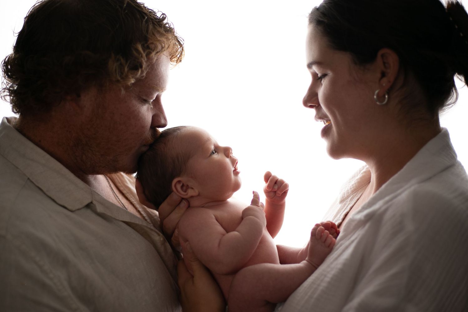 A silhouetted family shares tender moments with their baby against a bright white background.