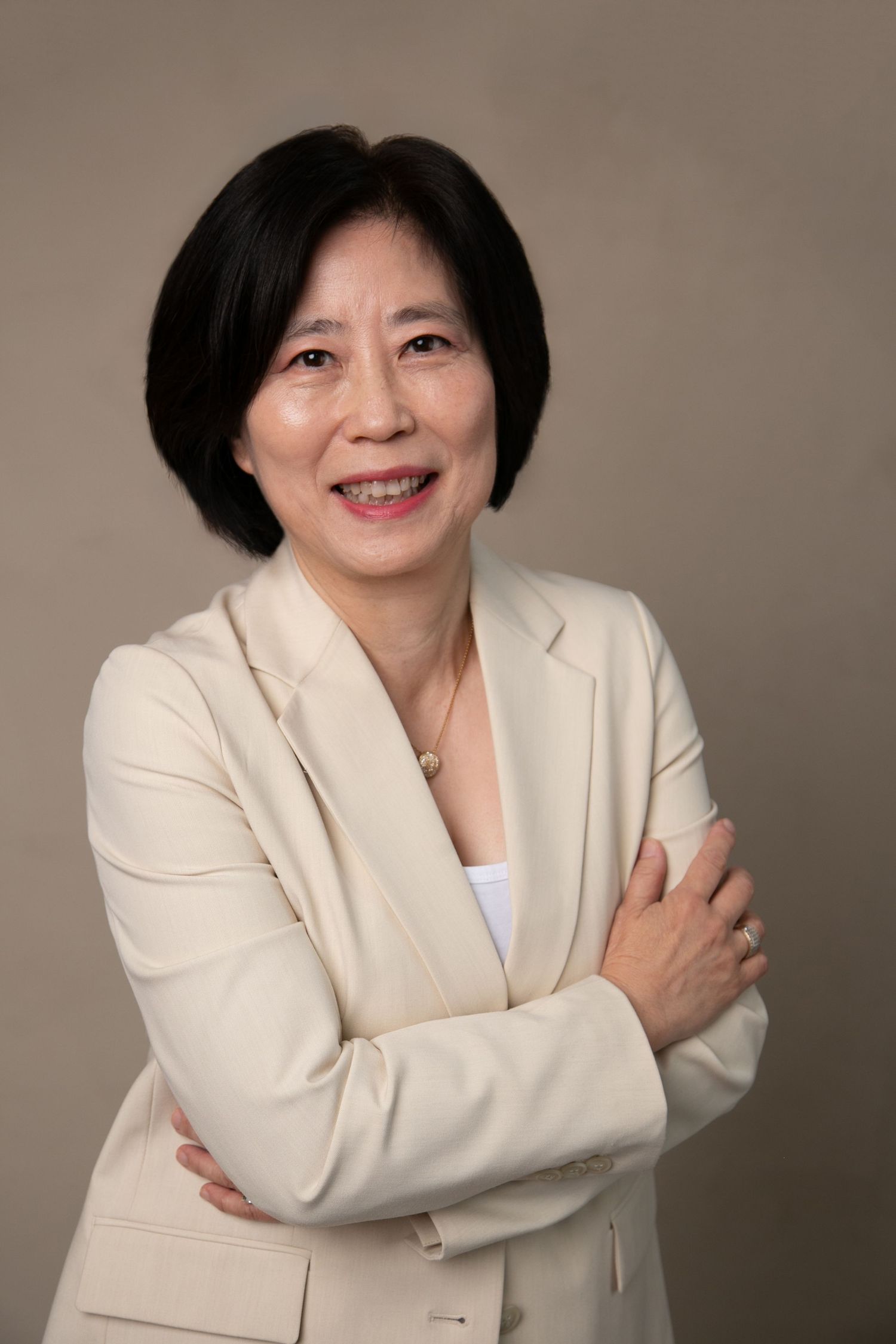Professional Asian woman in cream blazer smiling with arms crossed against neutral background.