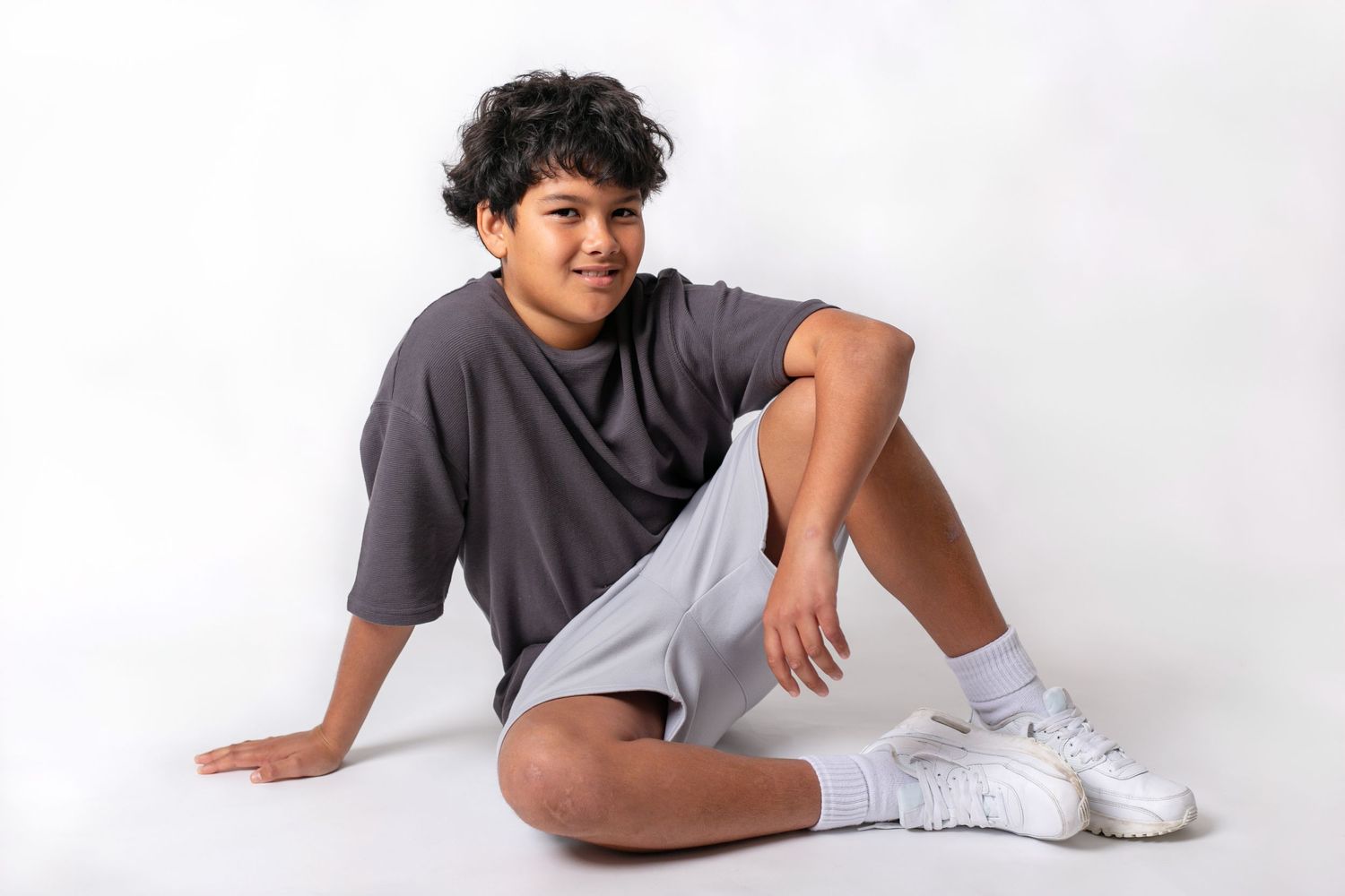 Person in gray shirt and white shorts reclines casually against white studio background.